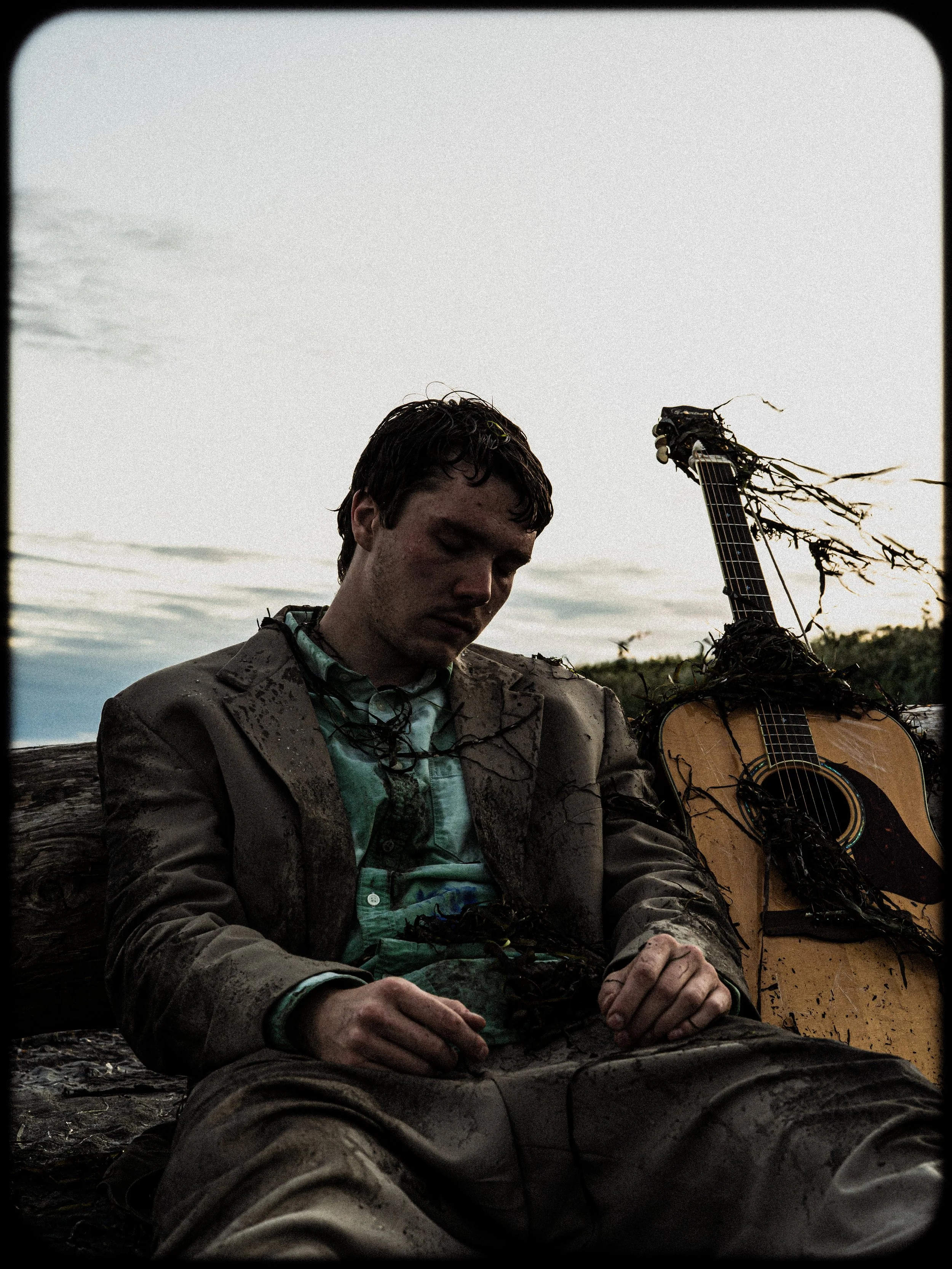 A young man in a muddy suit sits on a log with a battered guitar beside him, both covered in mud and vines, outdoors during a cloudy evening.