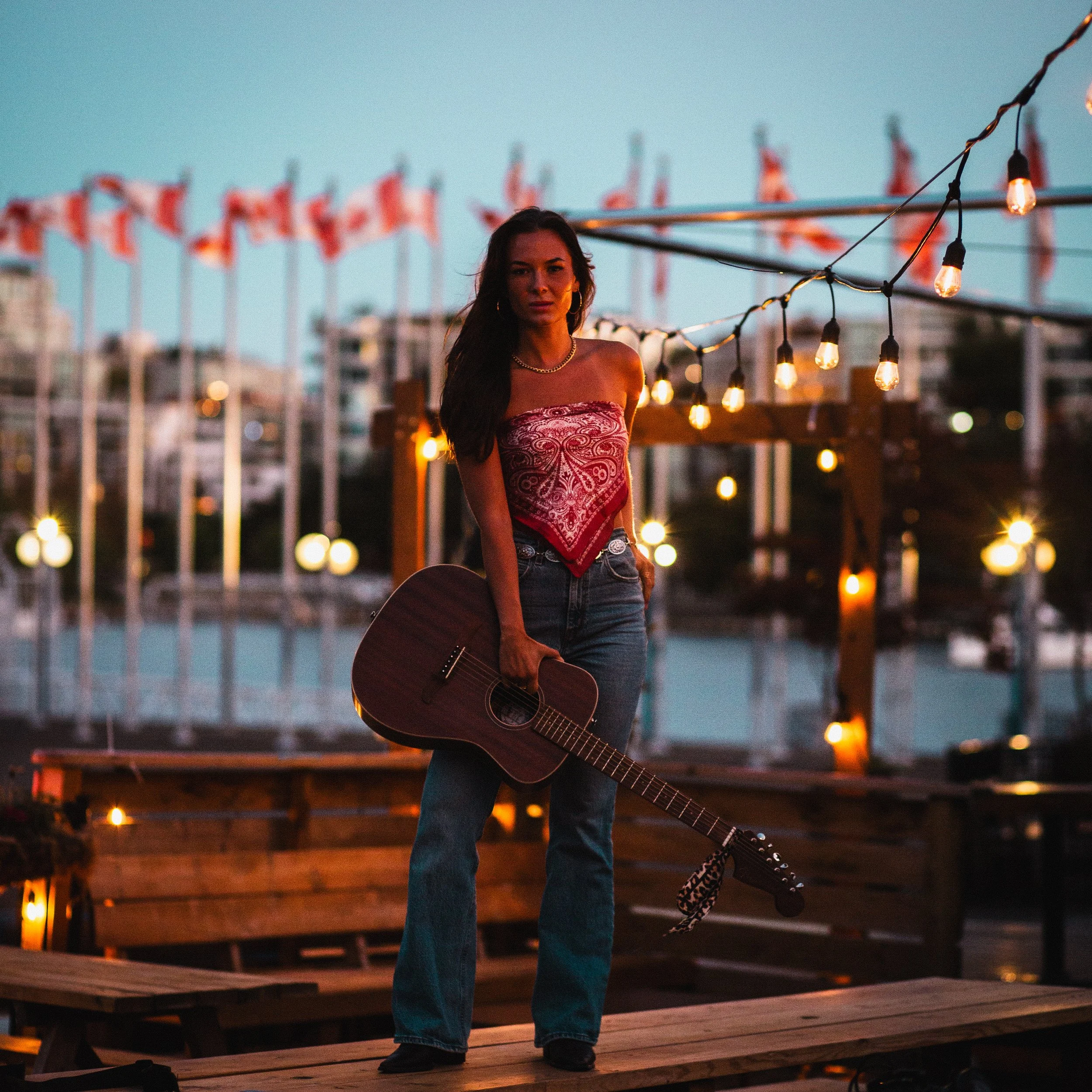 A woman standing on a wooden platform holding a guitar, with string lights overhead and a blurry cityscape in the background during dusk.