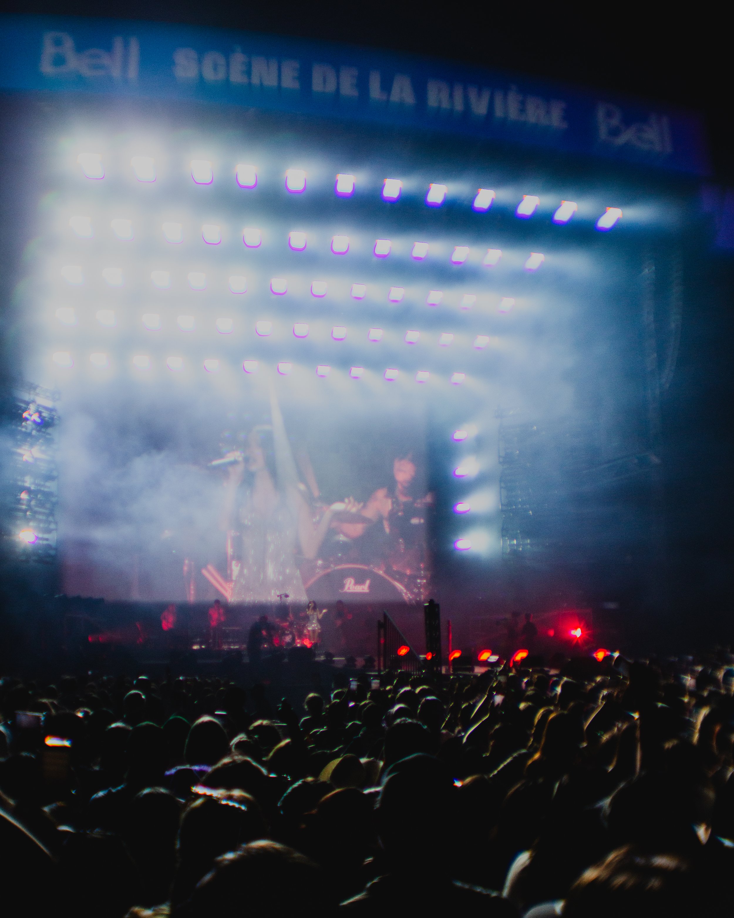 A concert stage with bright lights and smoke, viewed from the audience, with a crowd in front and a large screen in the background showing performers.