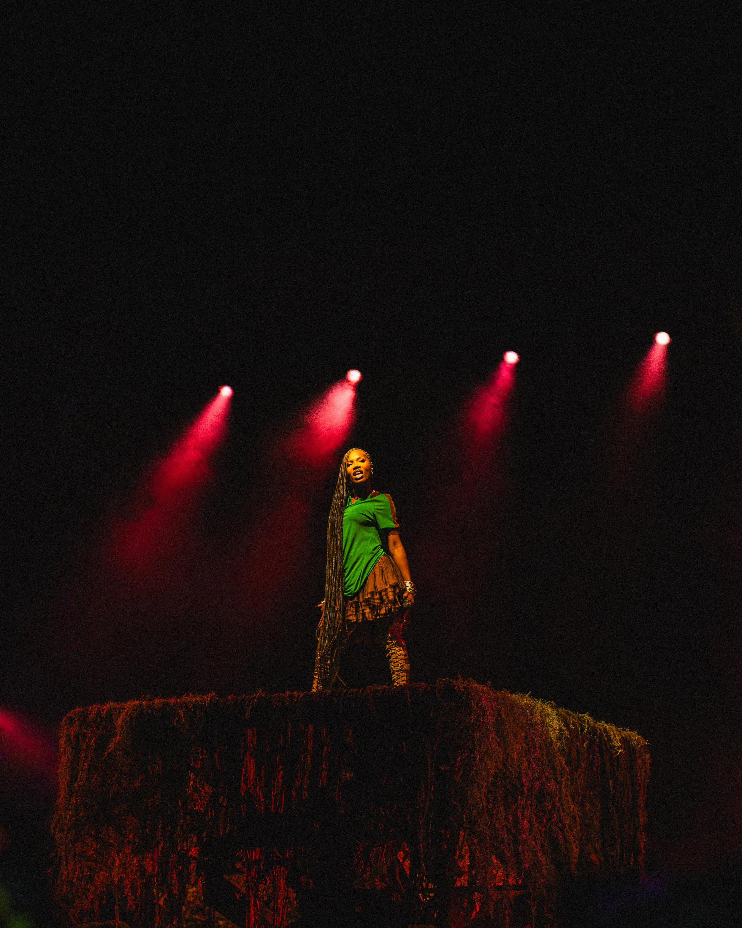 A woman on stage, wearing a green top and patterned pants, standing on a moss-covered platform with a dark background and red spotlights overhead.