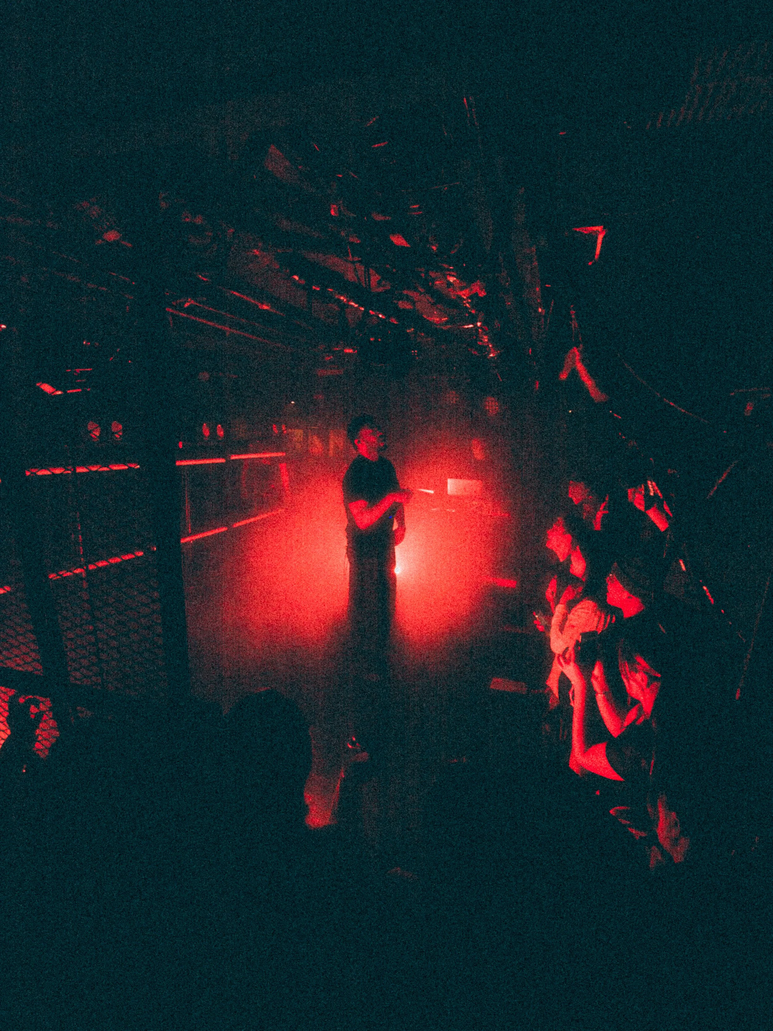Person standing on stage with red lighting, holding a microphone, with audience members seated in front.