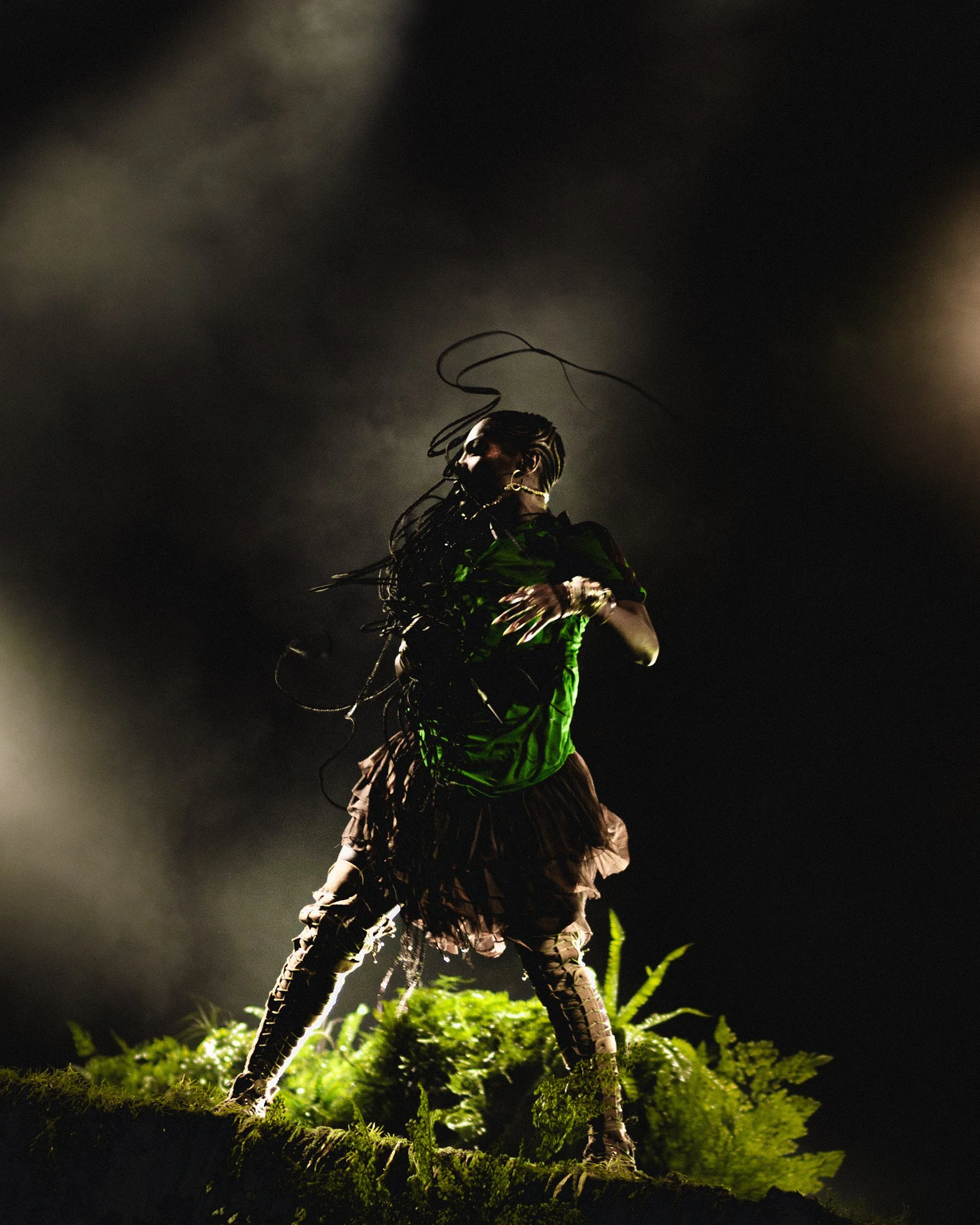 Performer on stage with dramatic lighting, wearing an elaborate costume with long braided hair, surrounded by foliage.