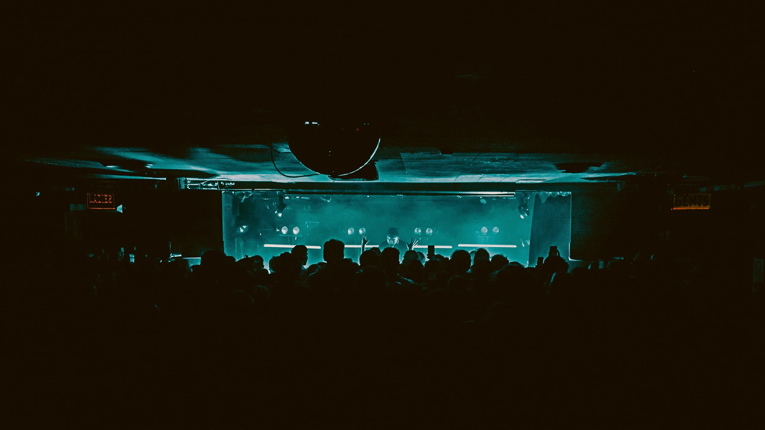 Dark indoor concert venue with a crowd facing a stage illuminated with blue-green lighting. The silhouette of the audience and several performers or DJs on stage are visible.
