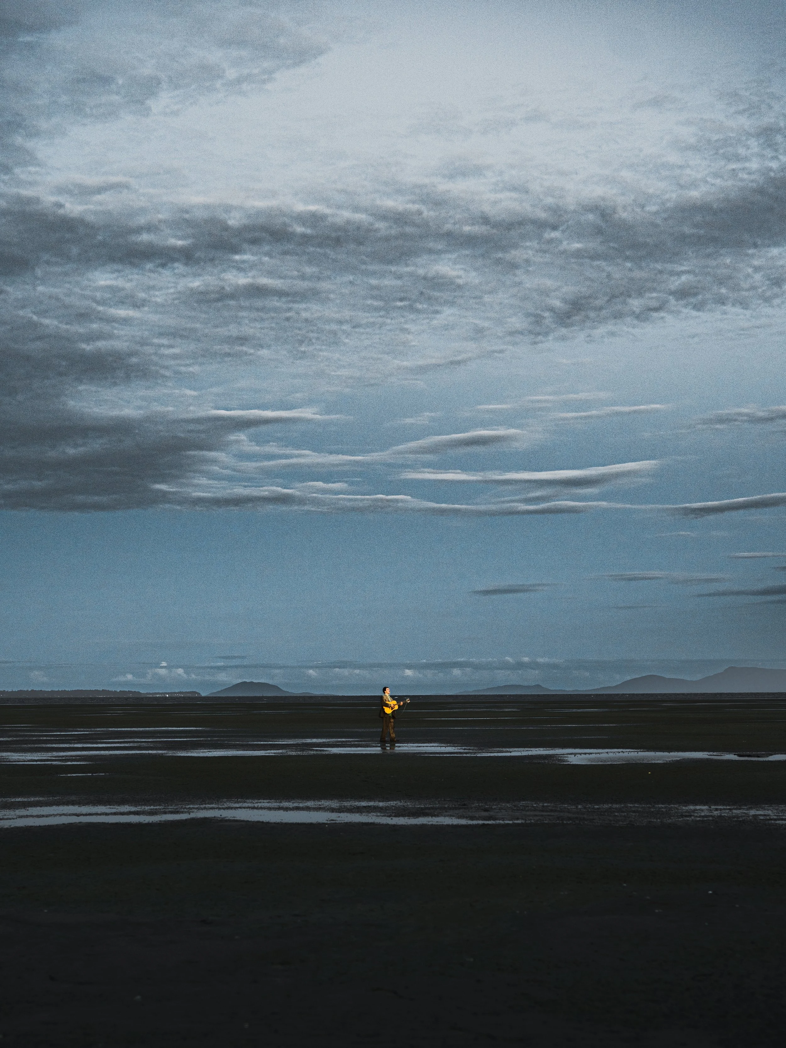 A person standing on a vast, partly flooded beach under a cloudy sky, holding an object, with mountains visible in the distant background.