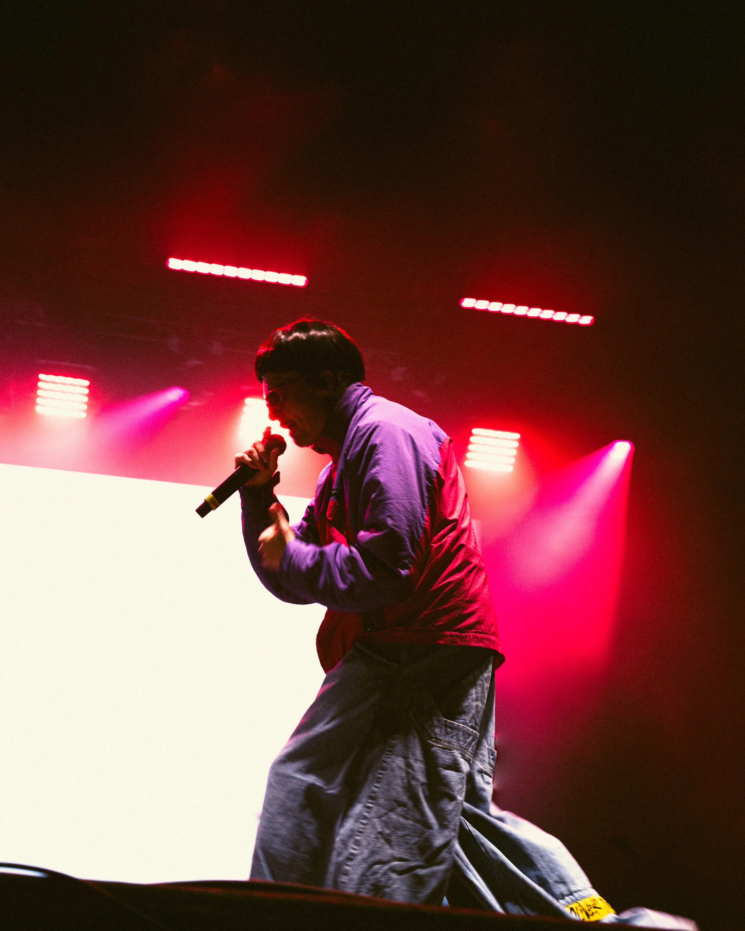 A performer singing into a microphone on stage with pink and red stage lights in the background.