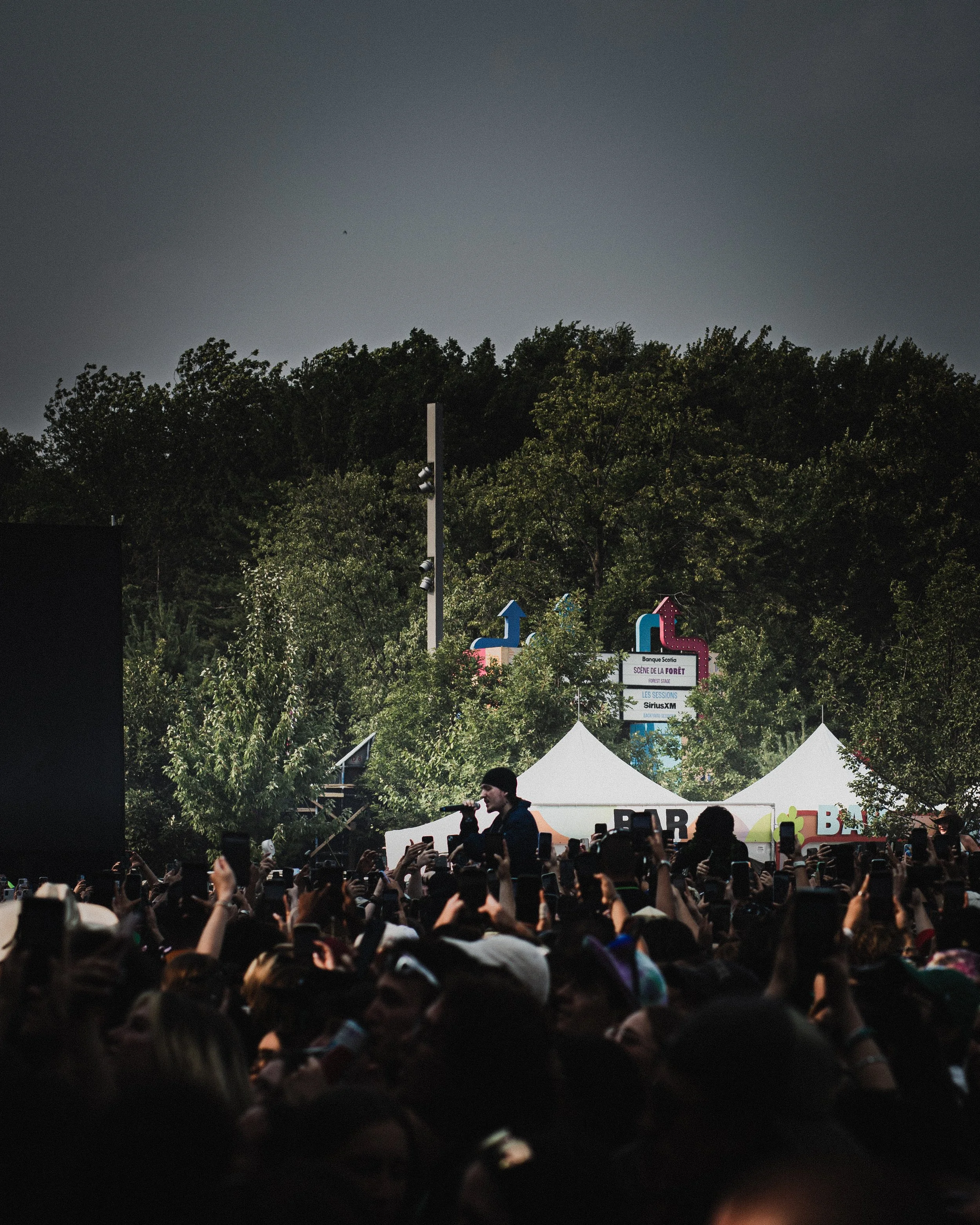 A live outdoor music concert with a crowd of people, a singer performing on stage, and trees in the background.