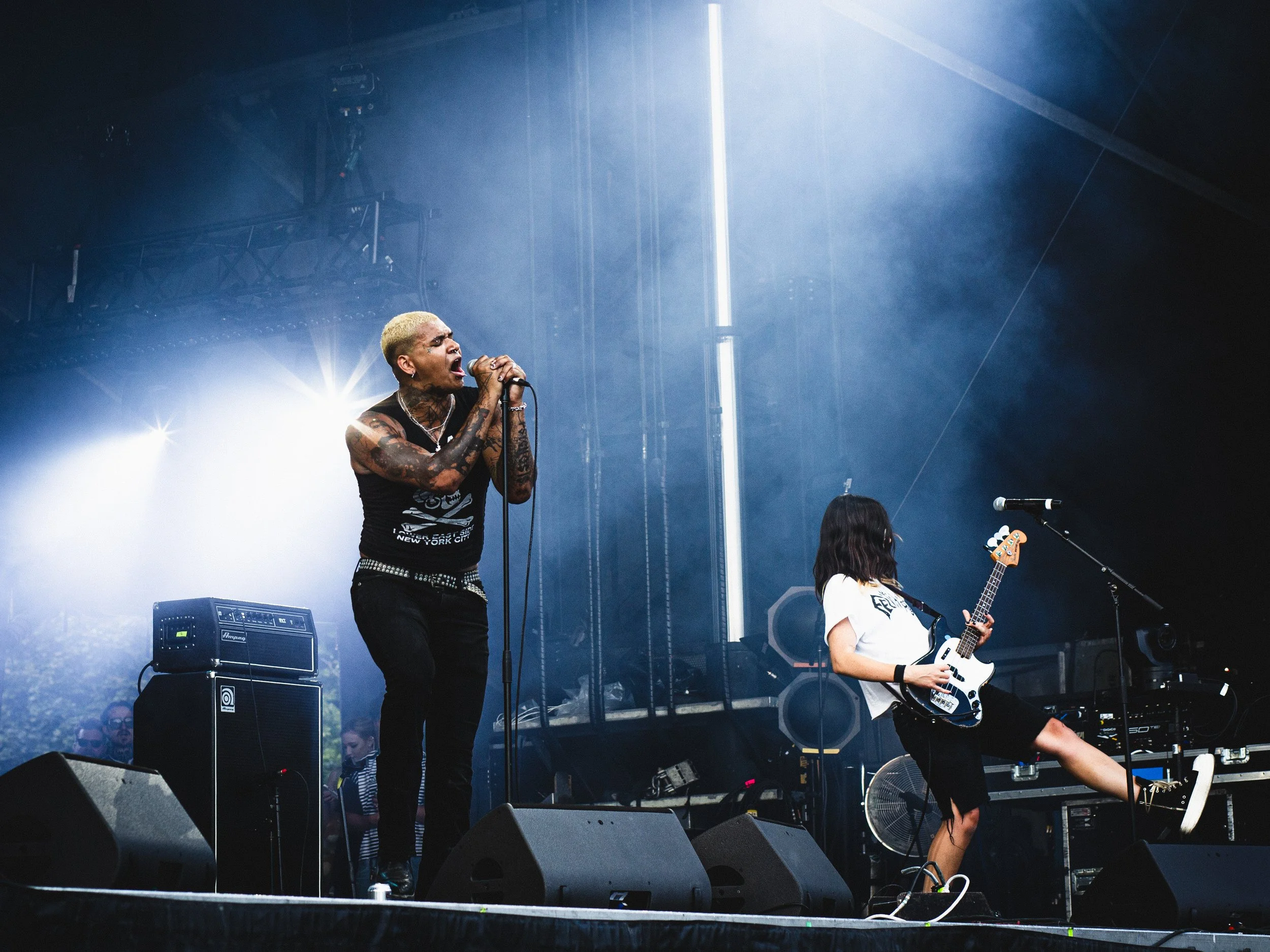 A band performing on stage with a lead singer singing into a microphone and a guitarist playing an electric guitar, with stage lighting and equipment visible.