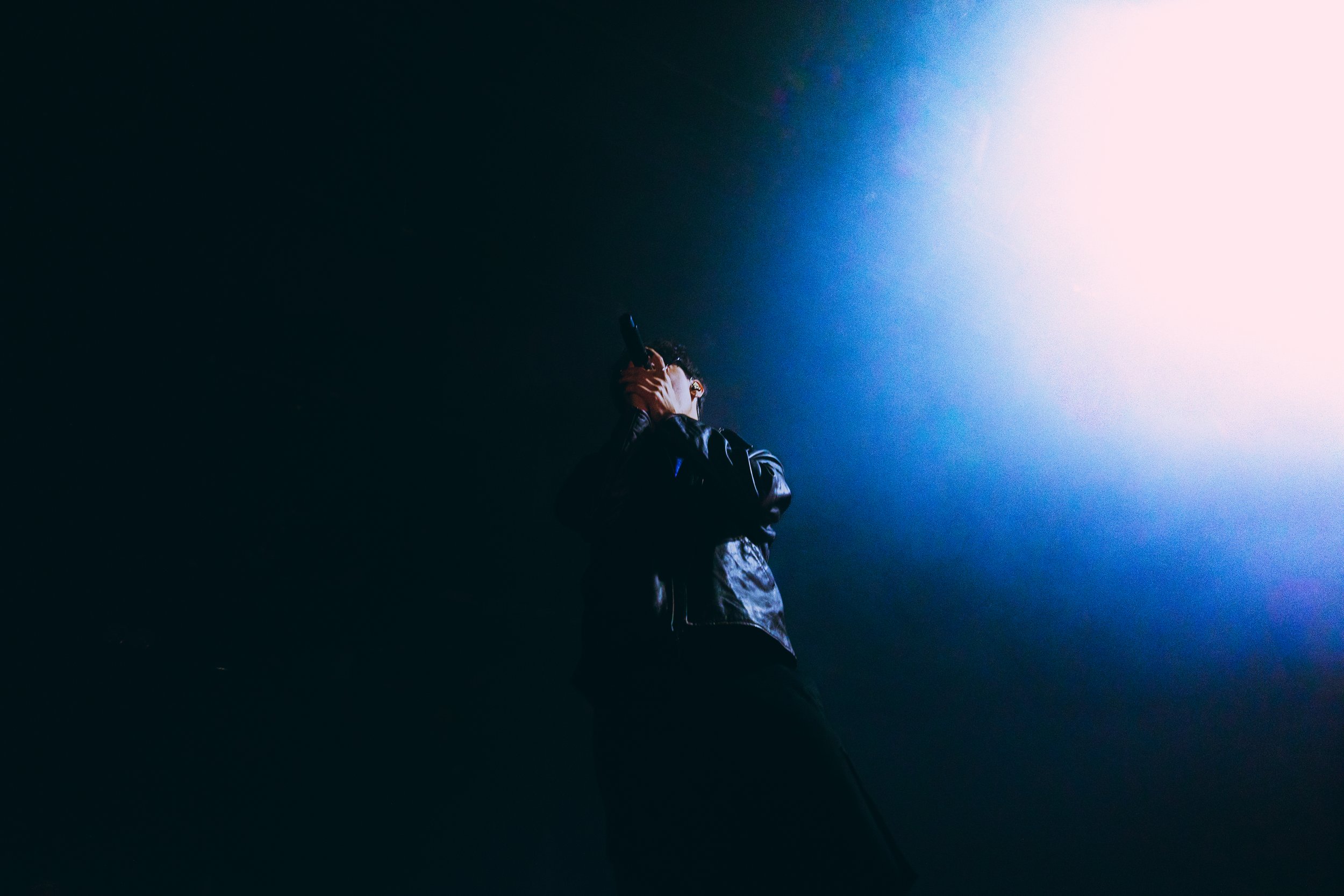 Person in leather jacket holding a microphone, singing or speaking, against a dark background with a bright light source on the right side.