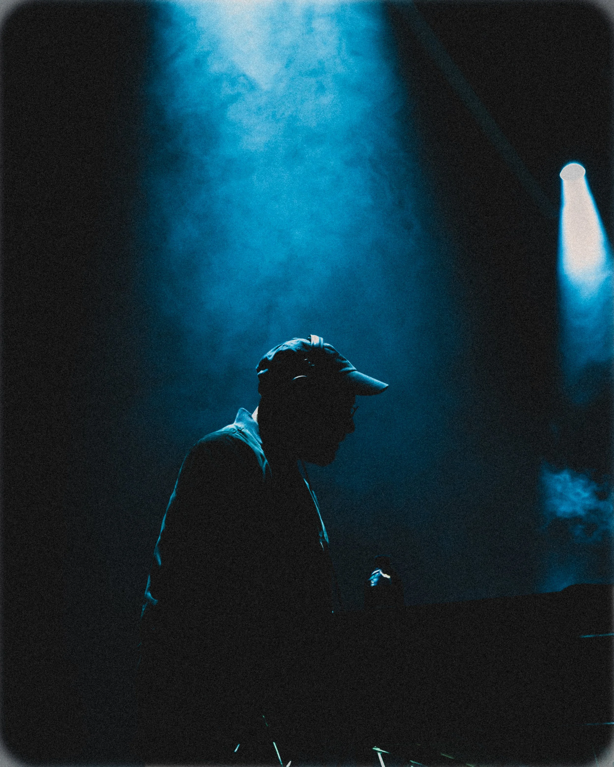 Silhouette of a DJ wearing a cap and glasses, standing behind turntables in a dark, blue-lit environment with fog and spotlight.
