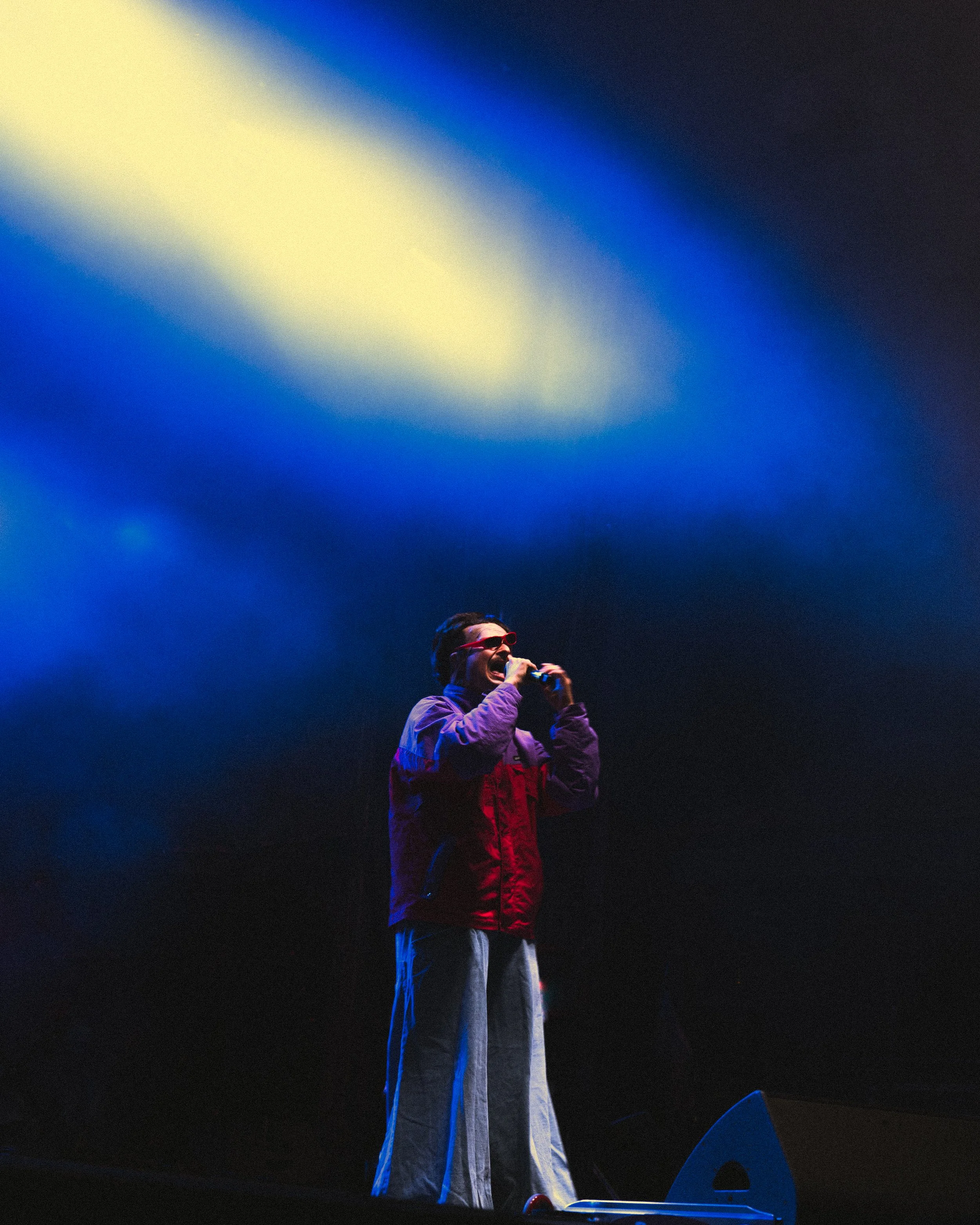 Performer singing on stage under colorful lights, wearing sunglasses, a red and purple jacket, and wide-legged pants.