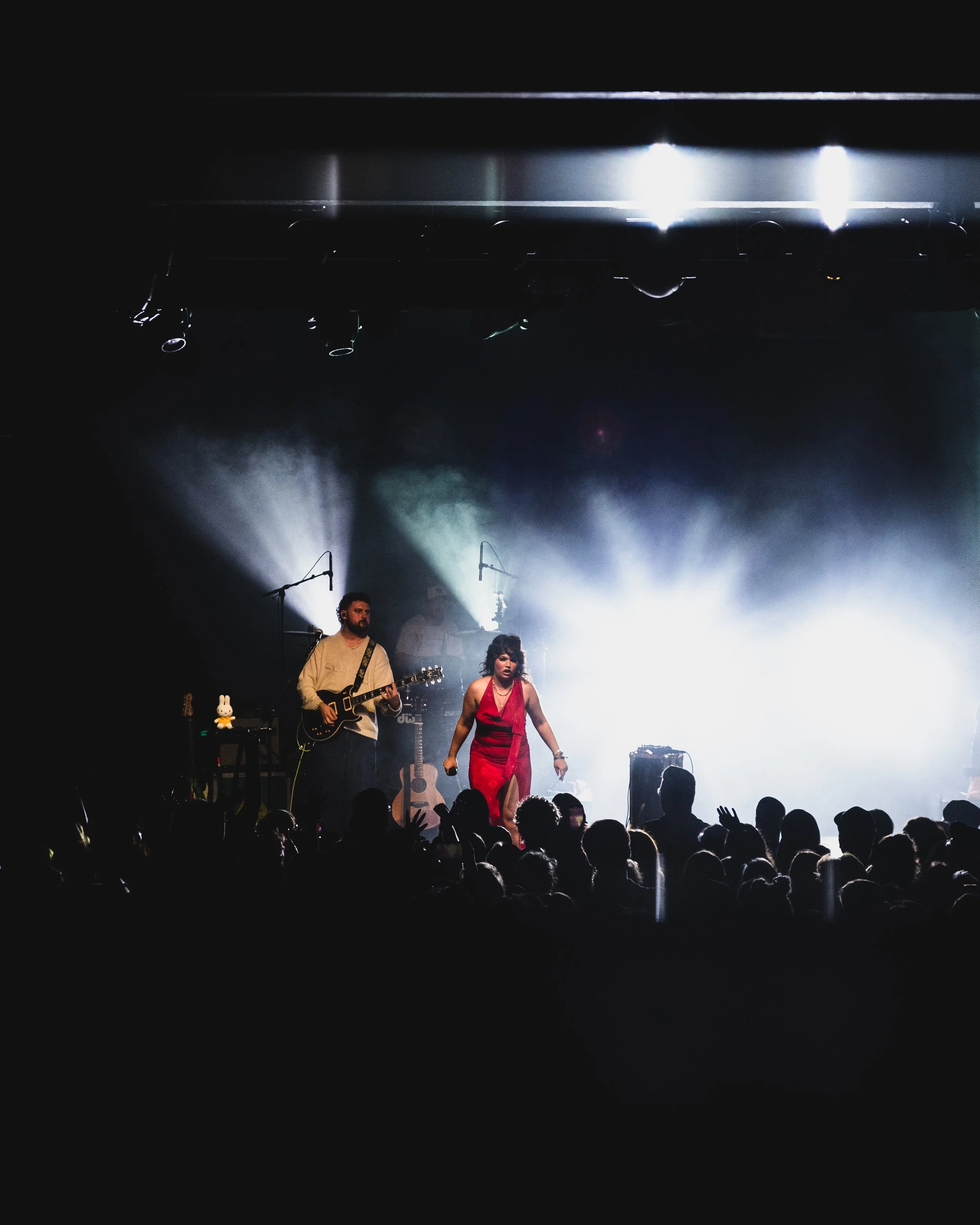 A concert stage with a female singer in a red dress performing, accompanied by a guitarist, with audience silhouettes in the foreground and bright stage lights behind.