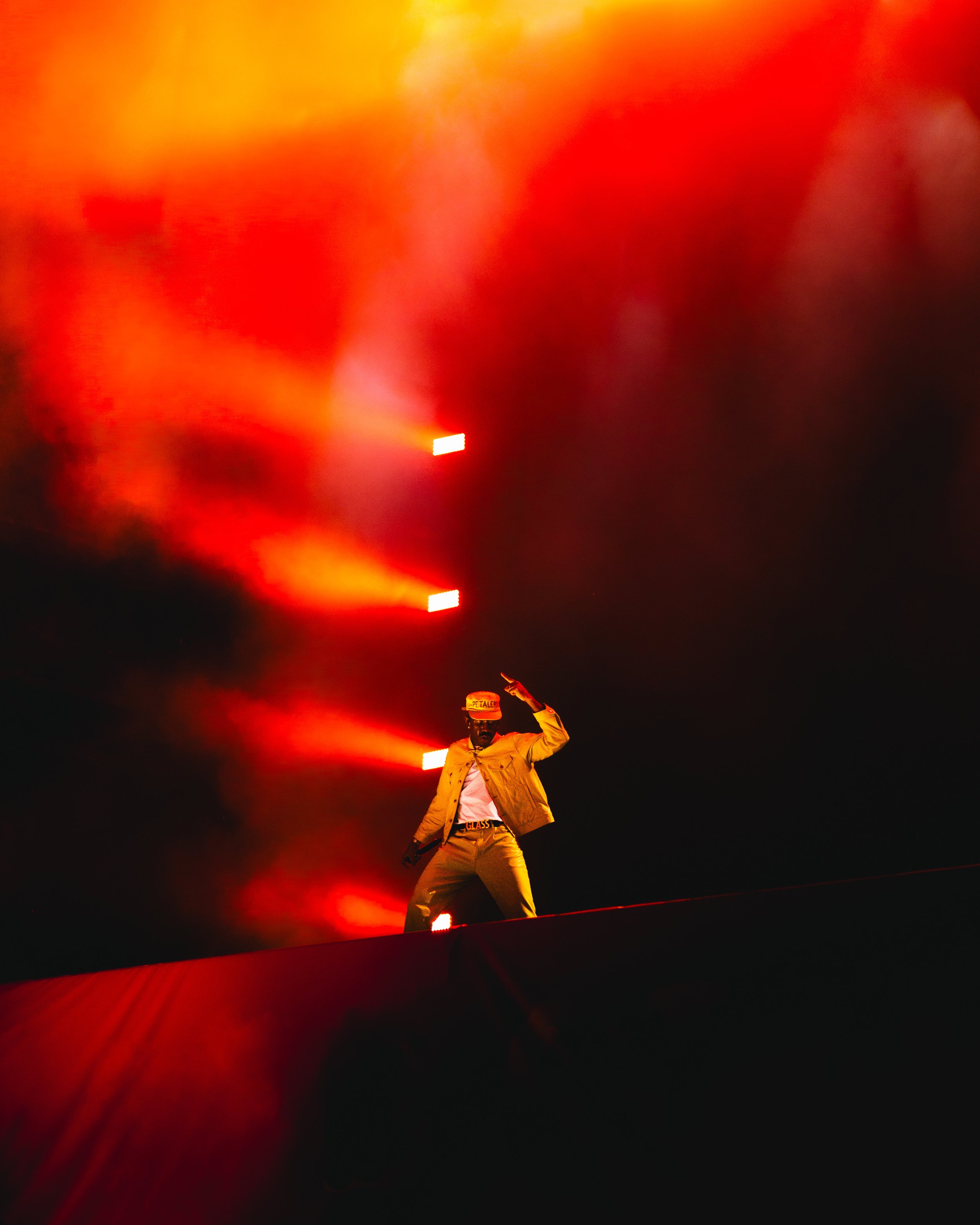 A person standing on stage illuminated by red stage lights, wearing a beige jacket, beige pants, a cap, and sunglasses, making a peace sign with one hand.