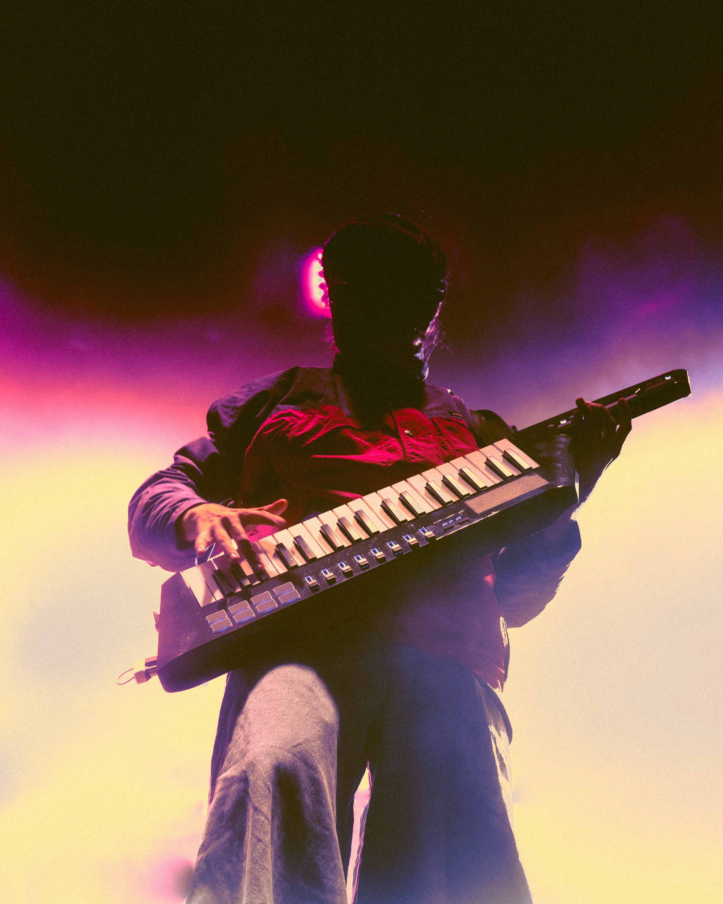 A person wearing a jacket playing a keyboard-like electronic instrument against a colorful stage background with purple and yellow lighting.
