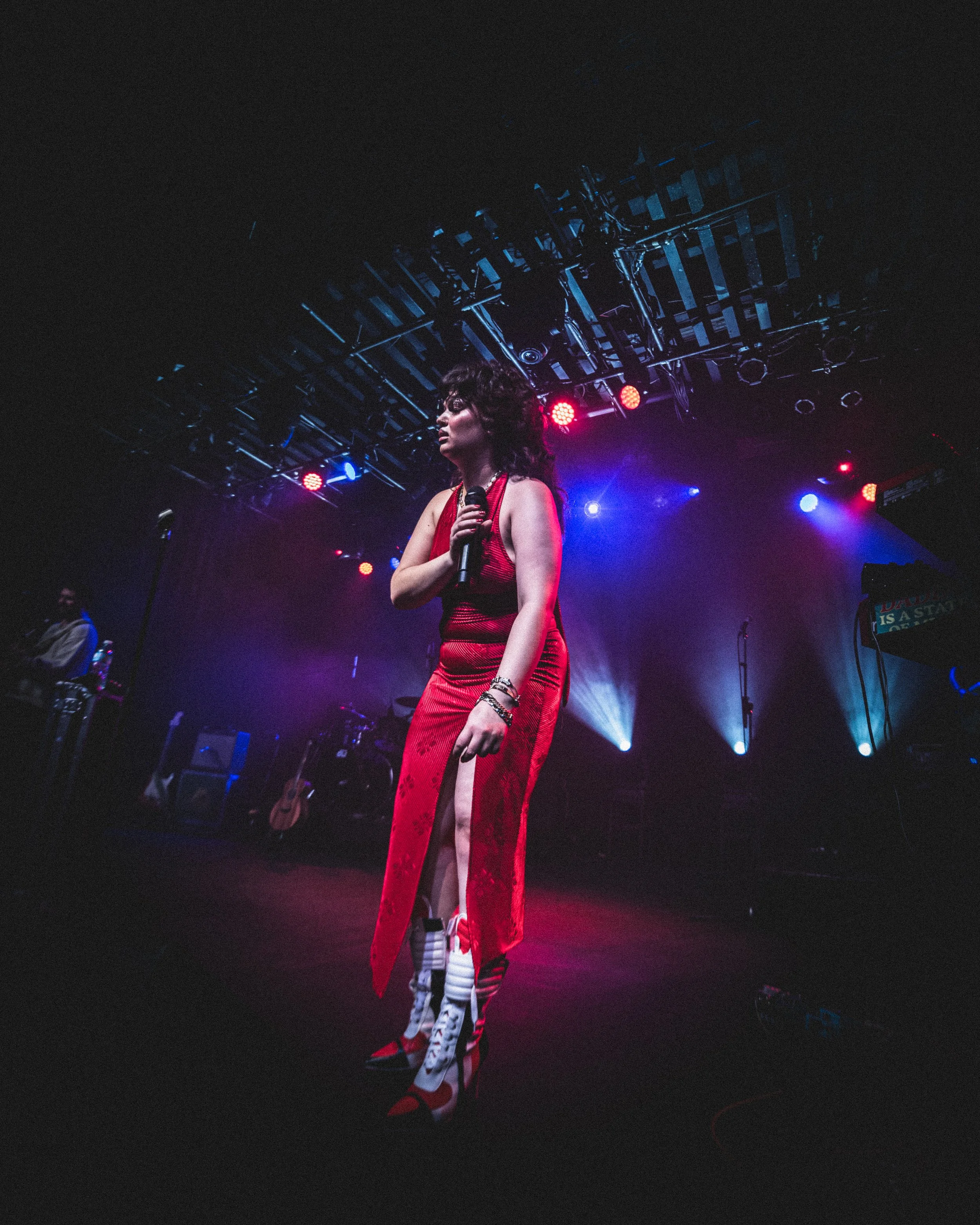 A woman in a red dress and high-heeled shoes holding a microphone on stage with colorful lights and musical instruments in the background.