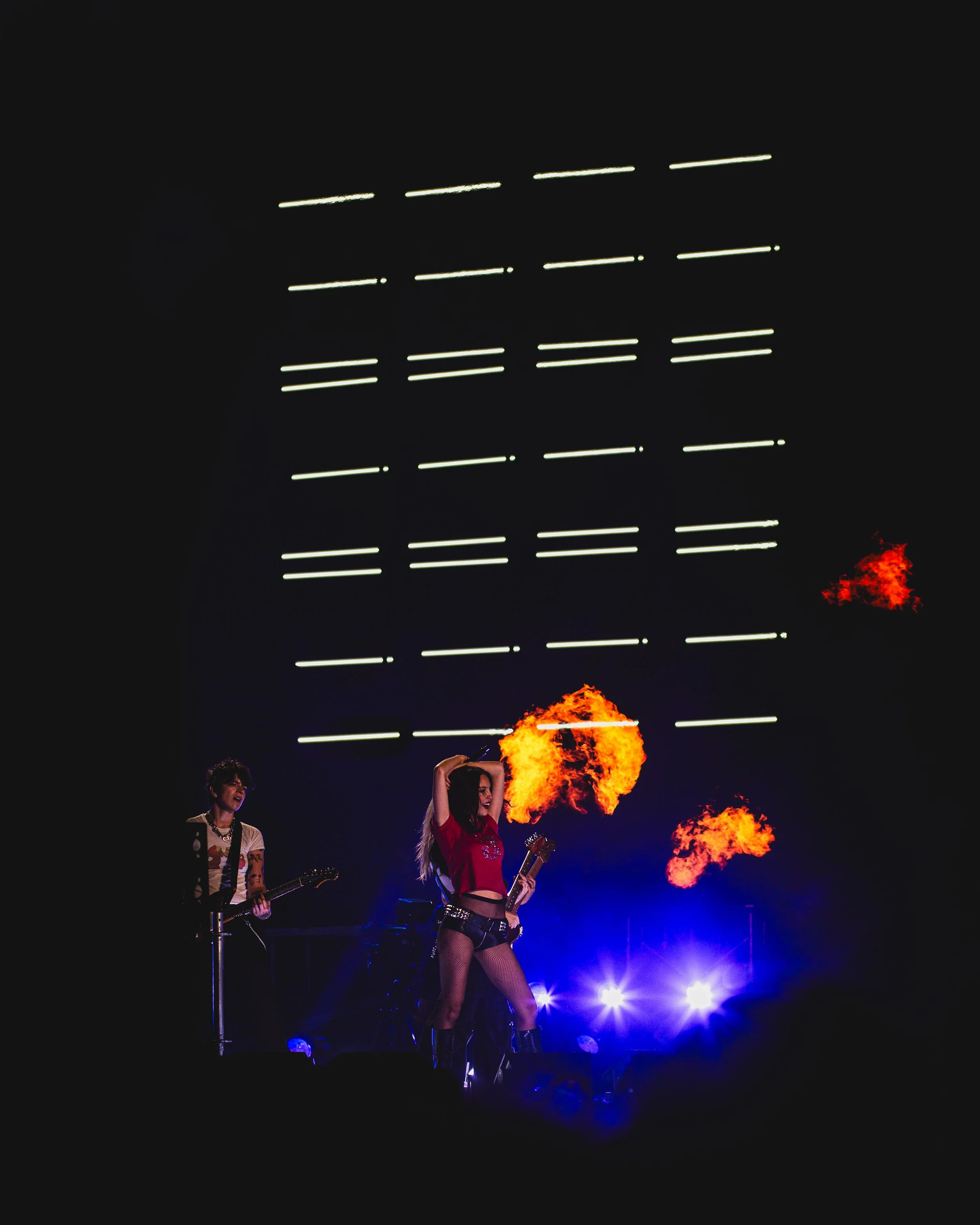 Performer with fiery torch on stage, illuminated by blue and white lights, with a musician playing guitar nearby, against a dark background with illuminated lines.