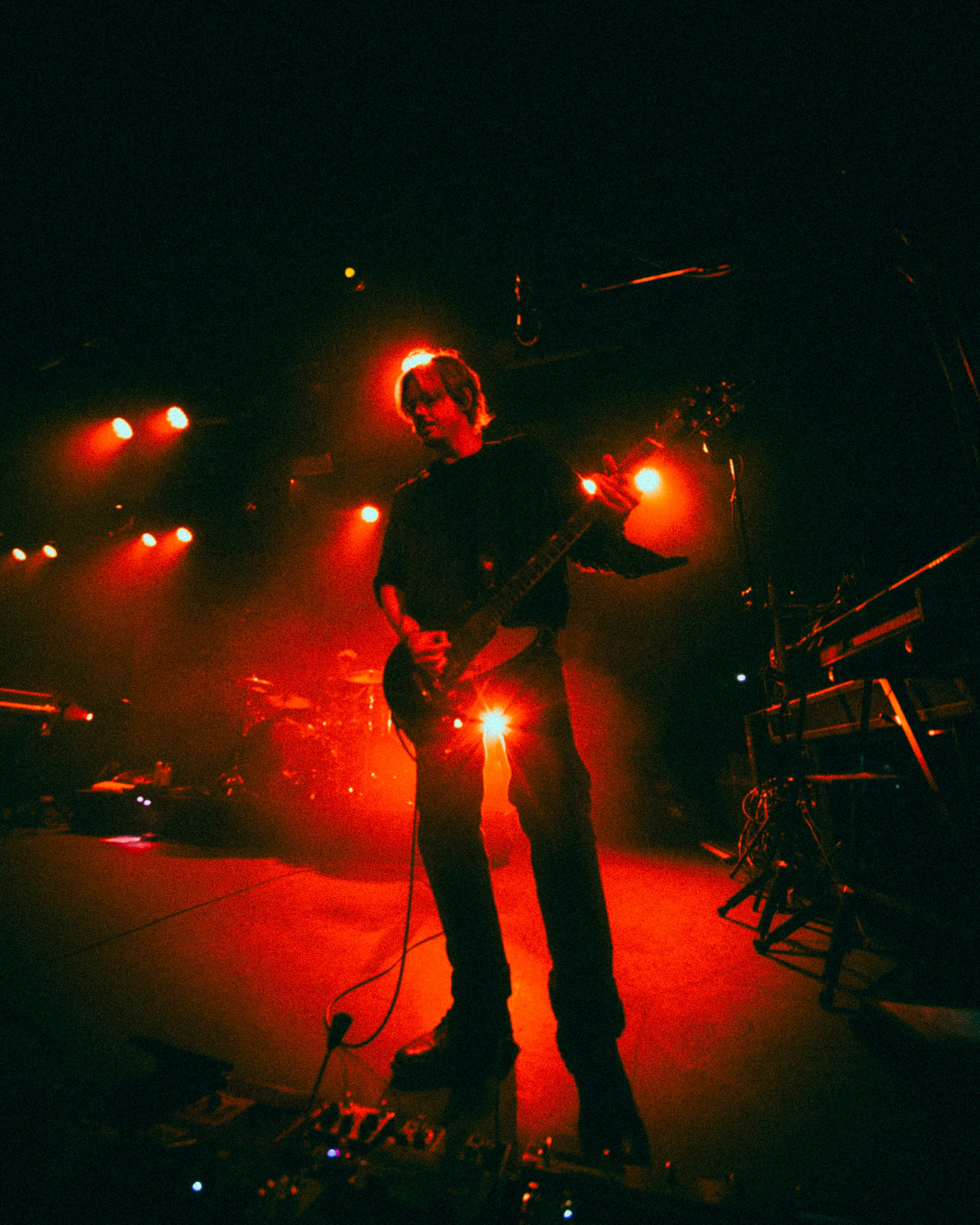 Musician playing electric guitar on stage with red lighting and sound equipment.