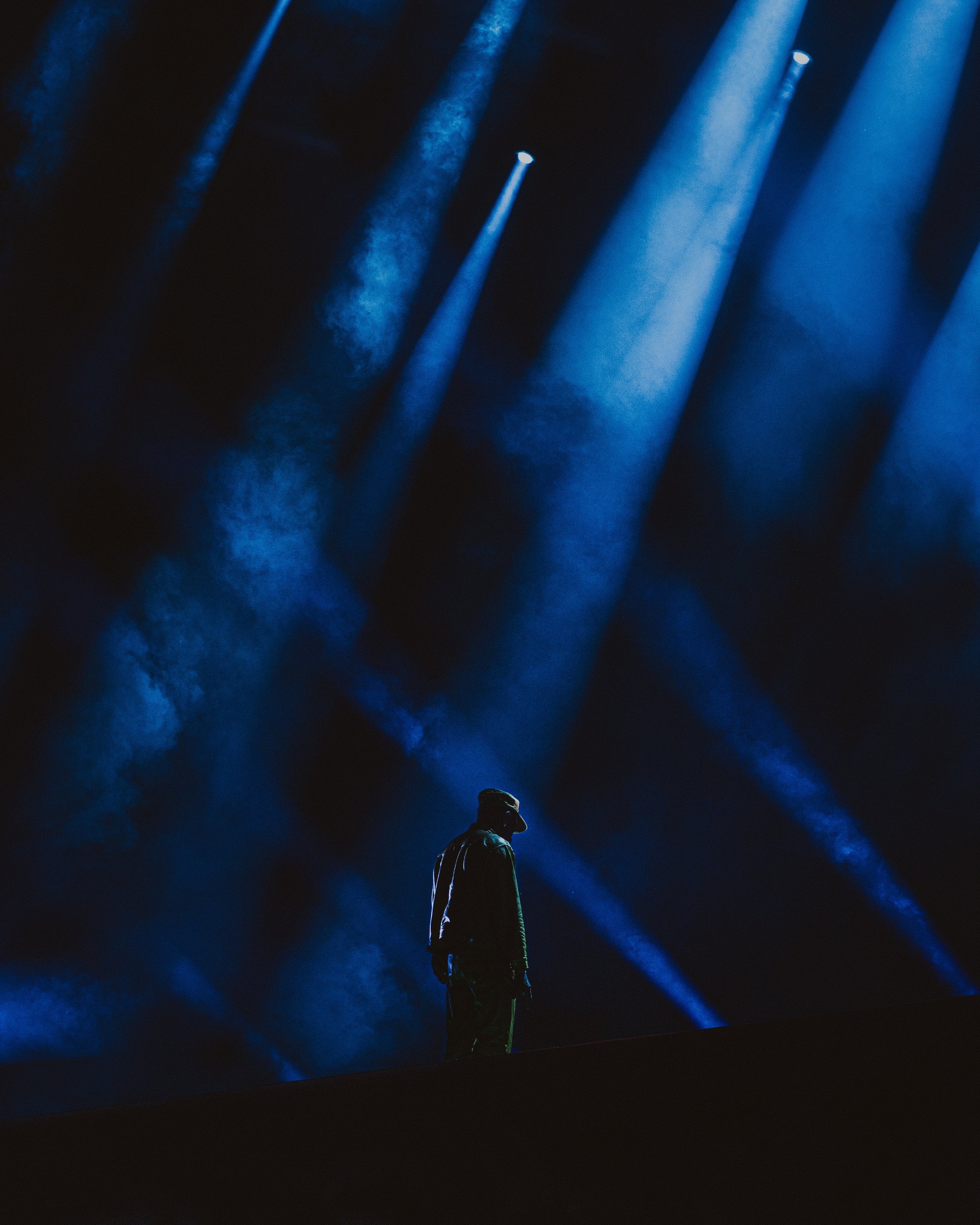 A person standing alone on a dark stage illuminated by blue spotlight beams creating an atmospheric scene.