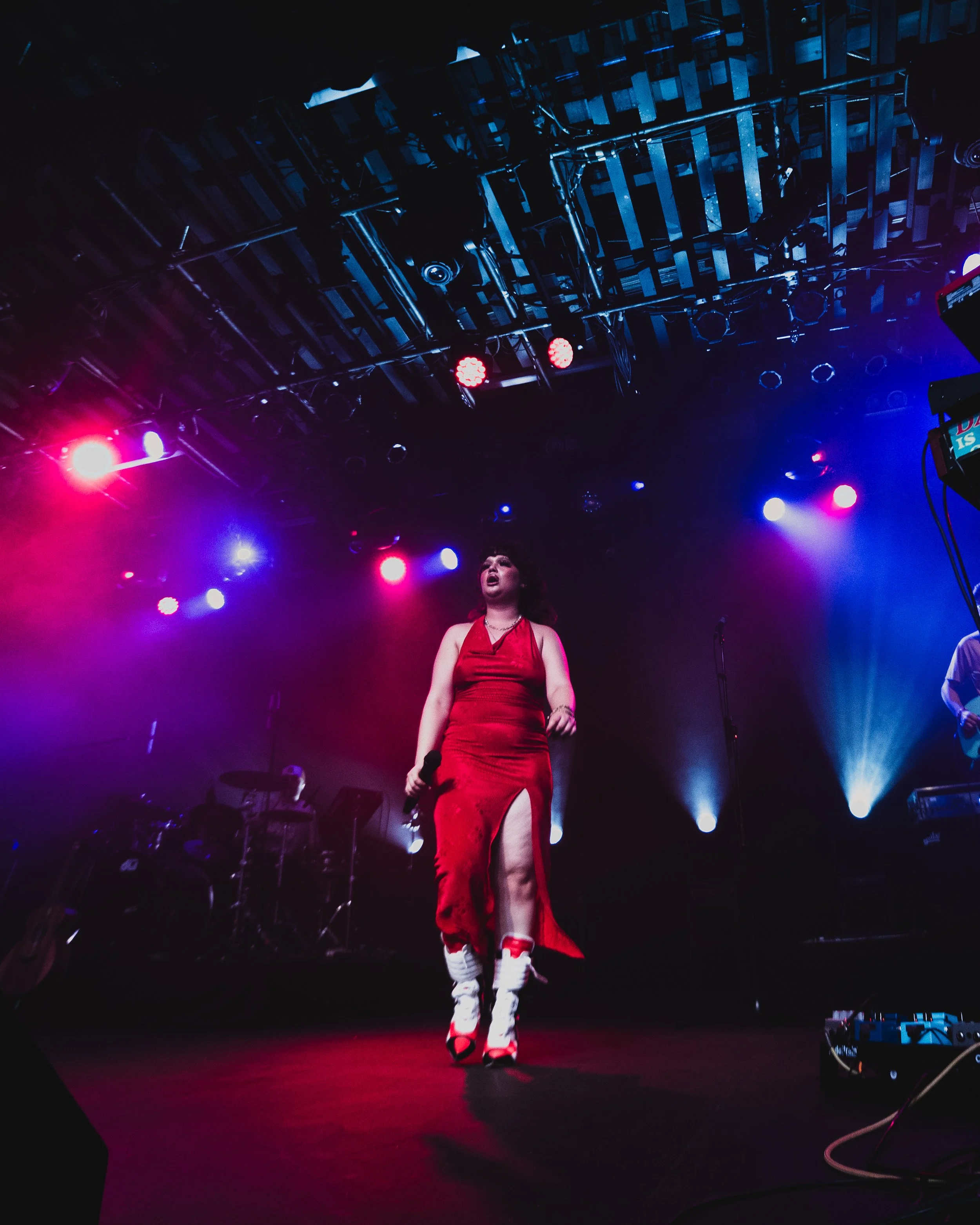 A female singer in a red dress with a slit walking on stage holding a microphone, with colorful stage lights illuminating the background.