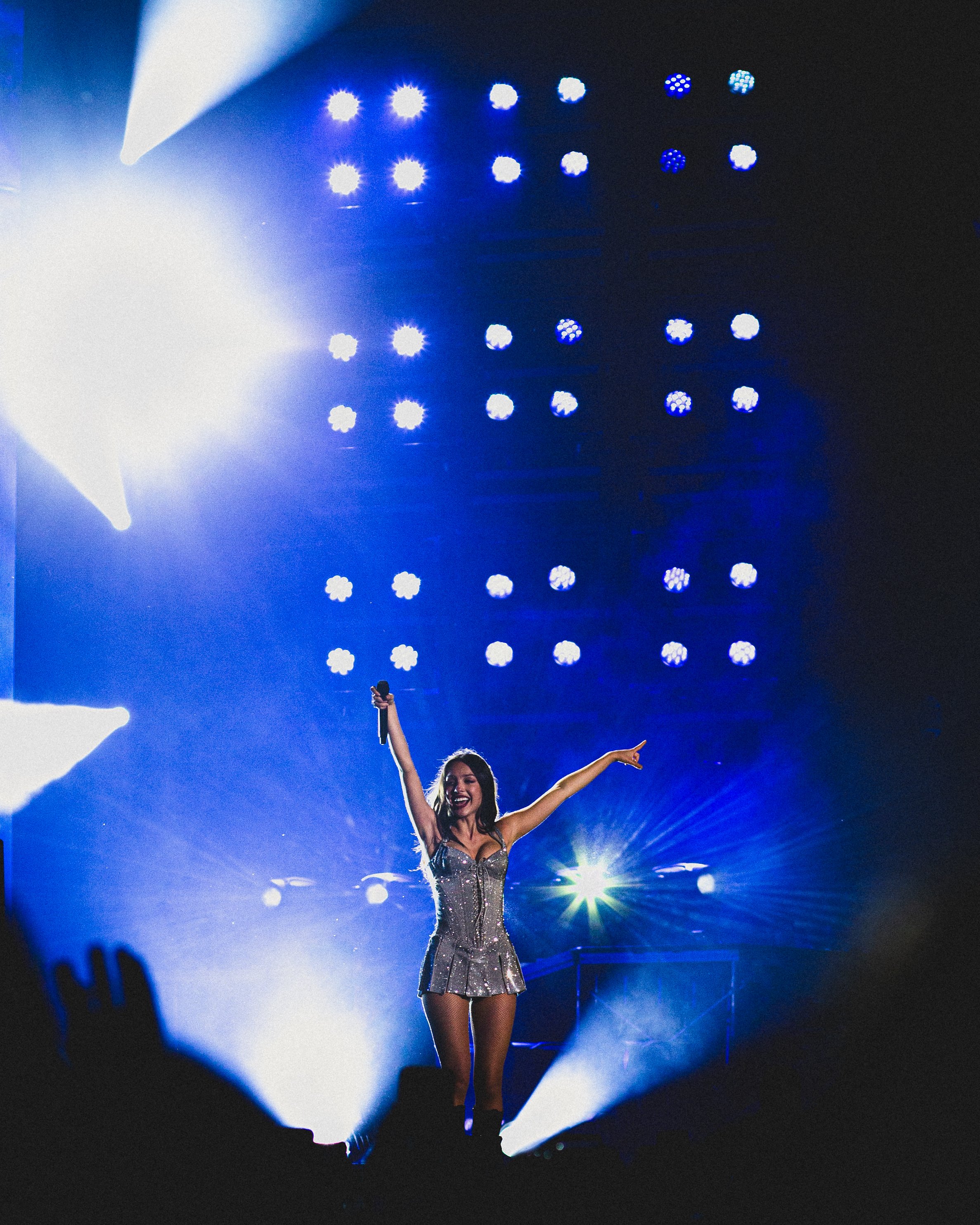 A female performer on stage with her arms raised, wearing a shiny silver dress, illuminated by bright stage lights in a concert setting.