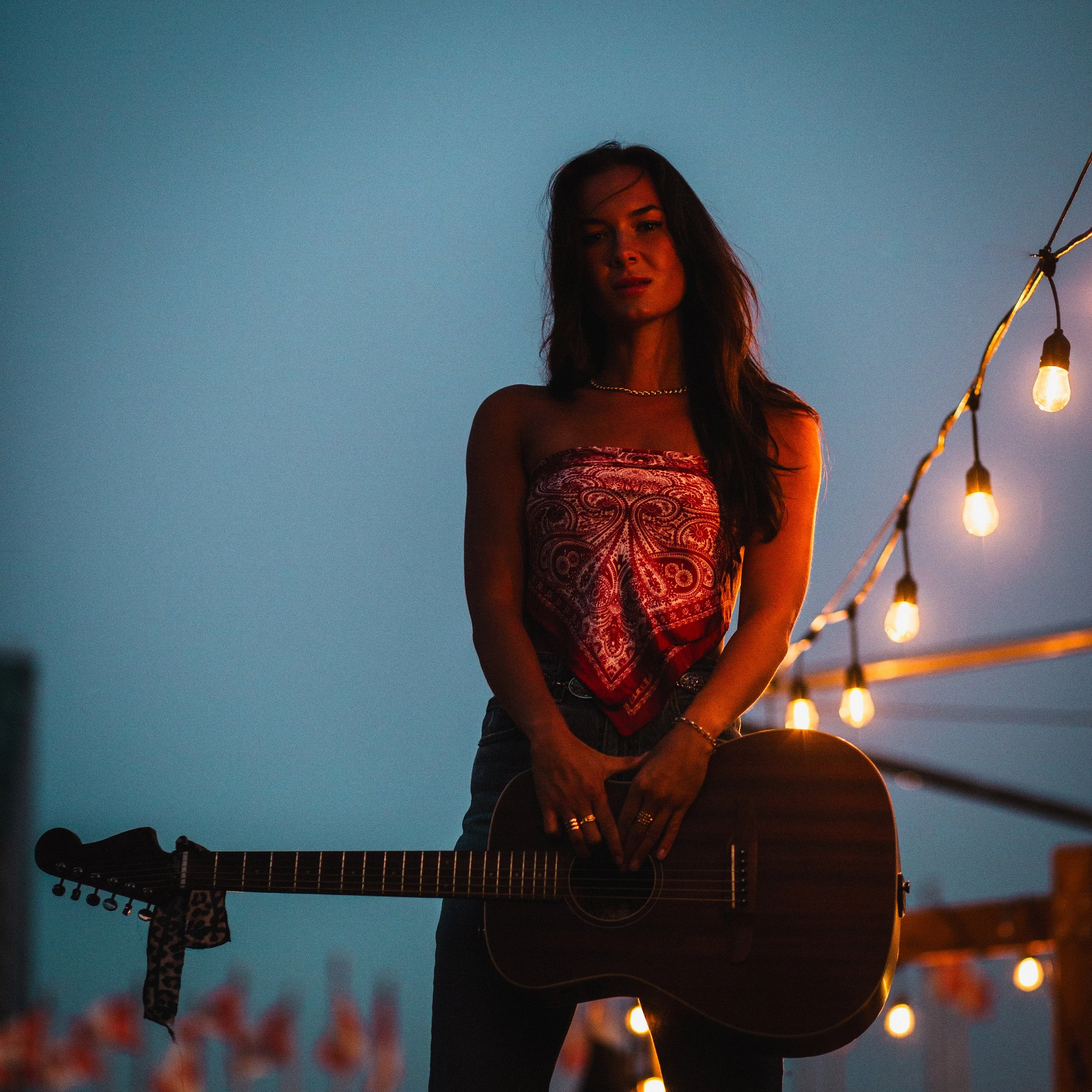 A woman with long hair standing outdoors at dusk, holding an acoustic guitar, with string lights hanging nearby.