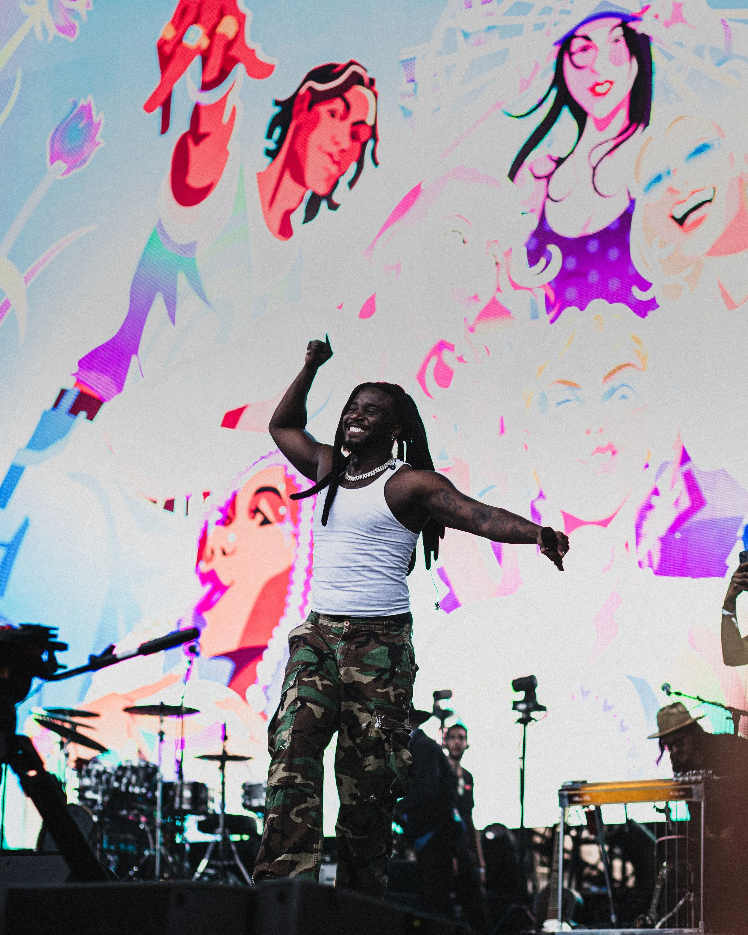 A man with long dreadlocks, wearing a white tank top, camouflage pants, and jewelry, dancing and smiling on stage with a colorful, animated cartoon mural behind him that includes multiple female faces and artistic designs.