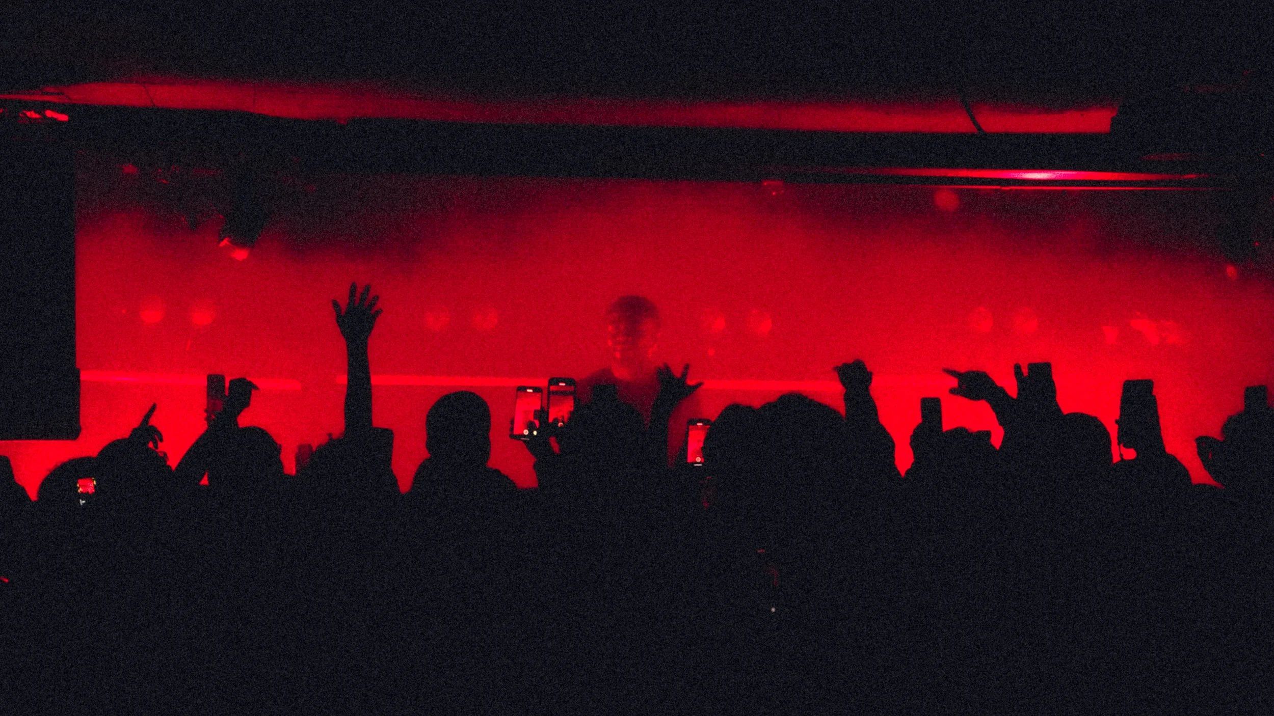 Silhouetted crowd with raised hands and smartphones in front of a red-lit DJ or performer at a nightclub or concert.