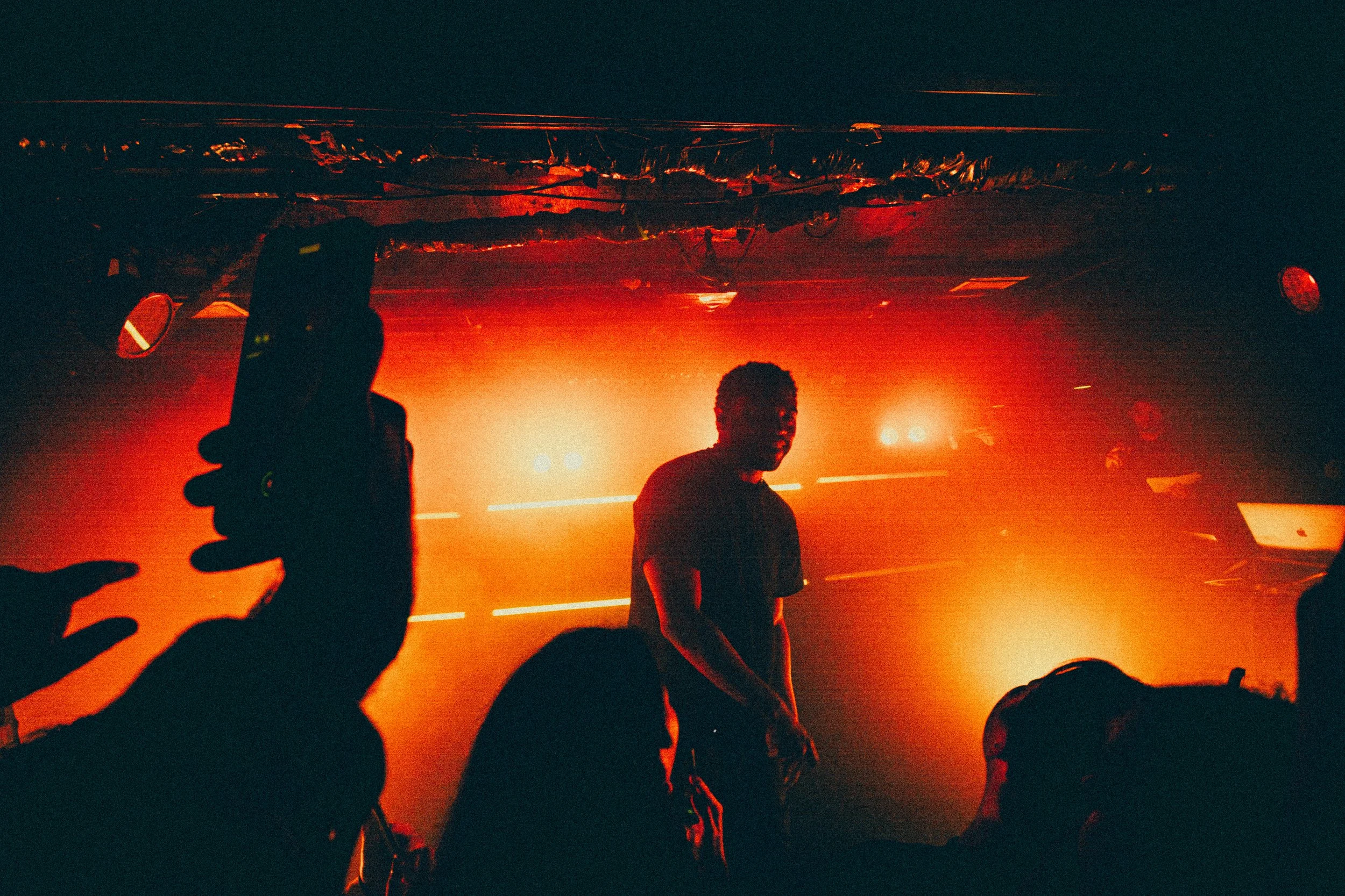 A DJ performing on stage with red lighting, surrounded by silhouettes of people at a nightclub or concert.
