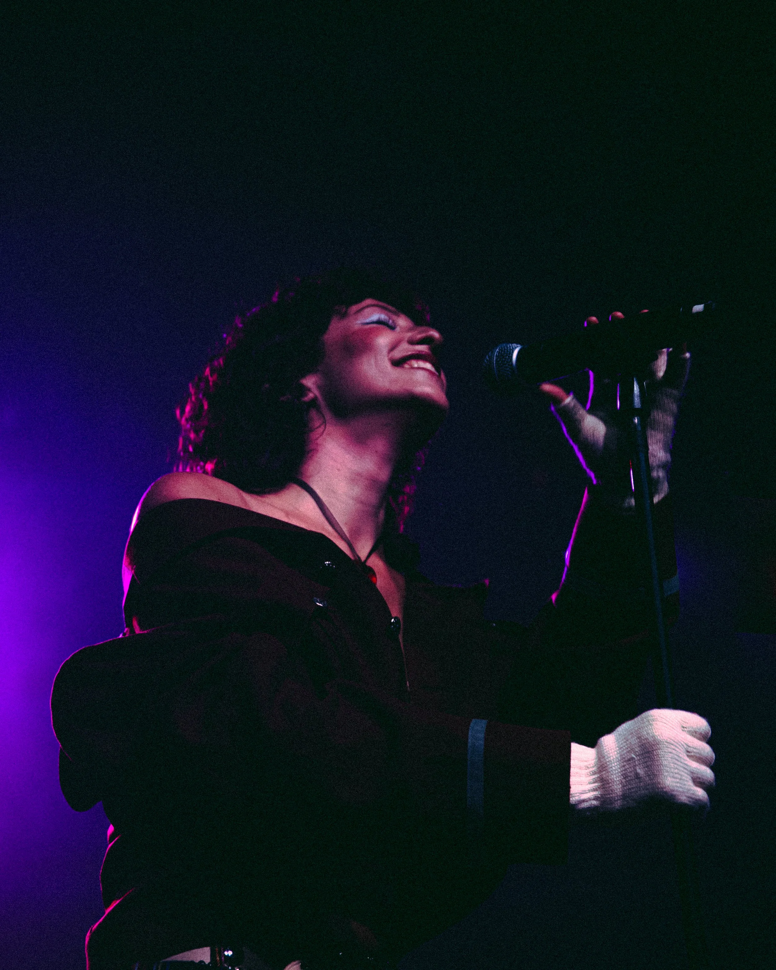A woman singing into a microphone on stage, smiling, with purple stage lighting.