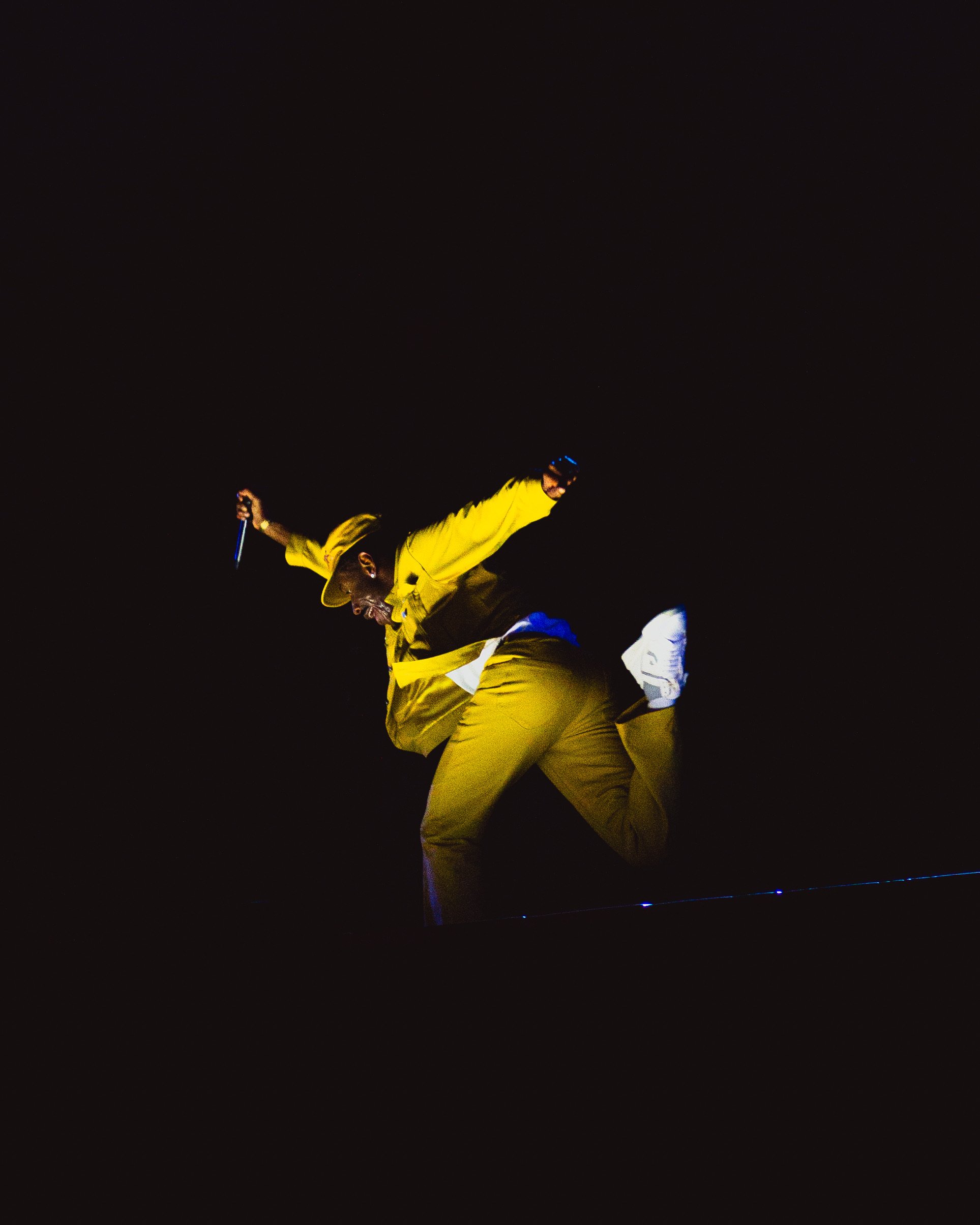 A man in a yellow suit and white sneakers jumping on a dark stage, holding a microphone in one hand.