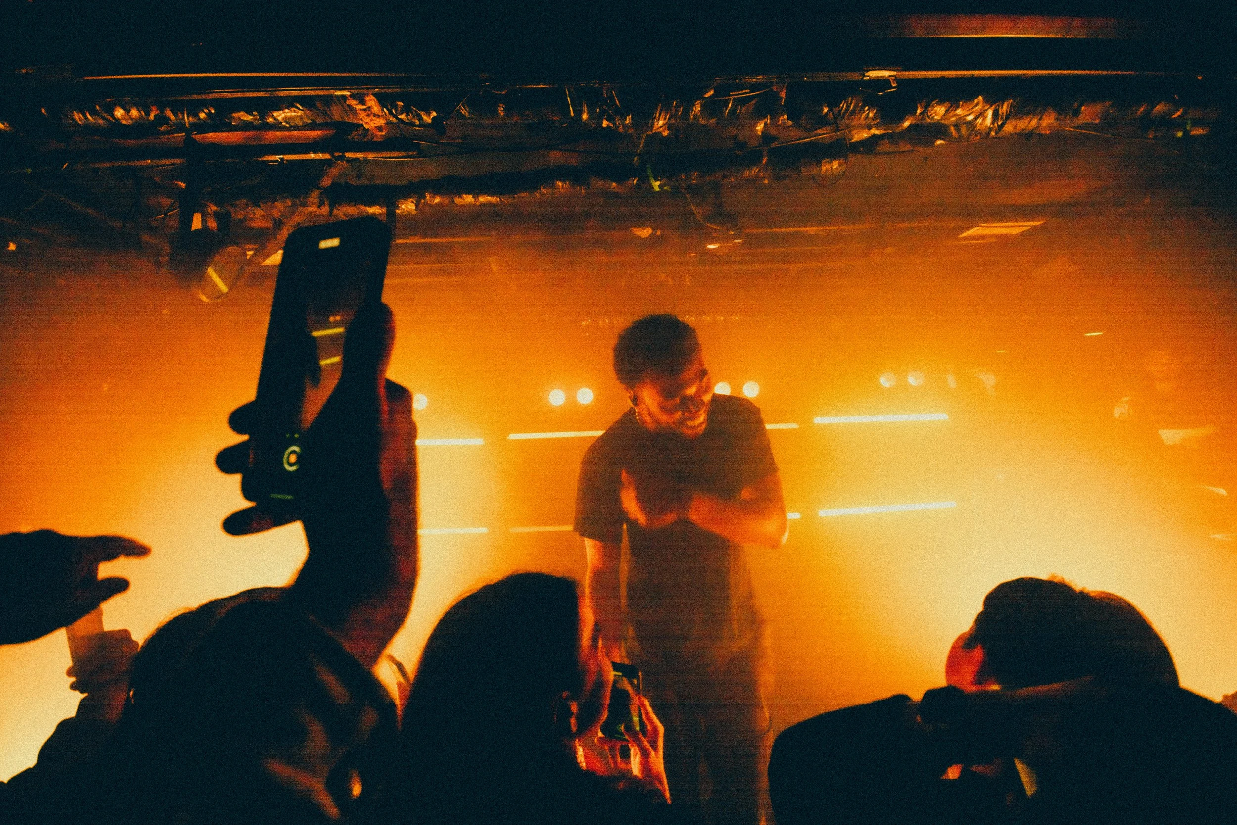 A performer on stage, smiling and engaging with the audience, with warm orange lighting and an audience member taking a photo with a smartphone.
