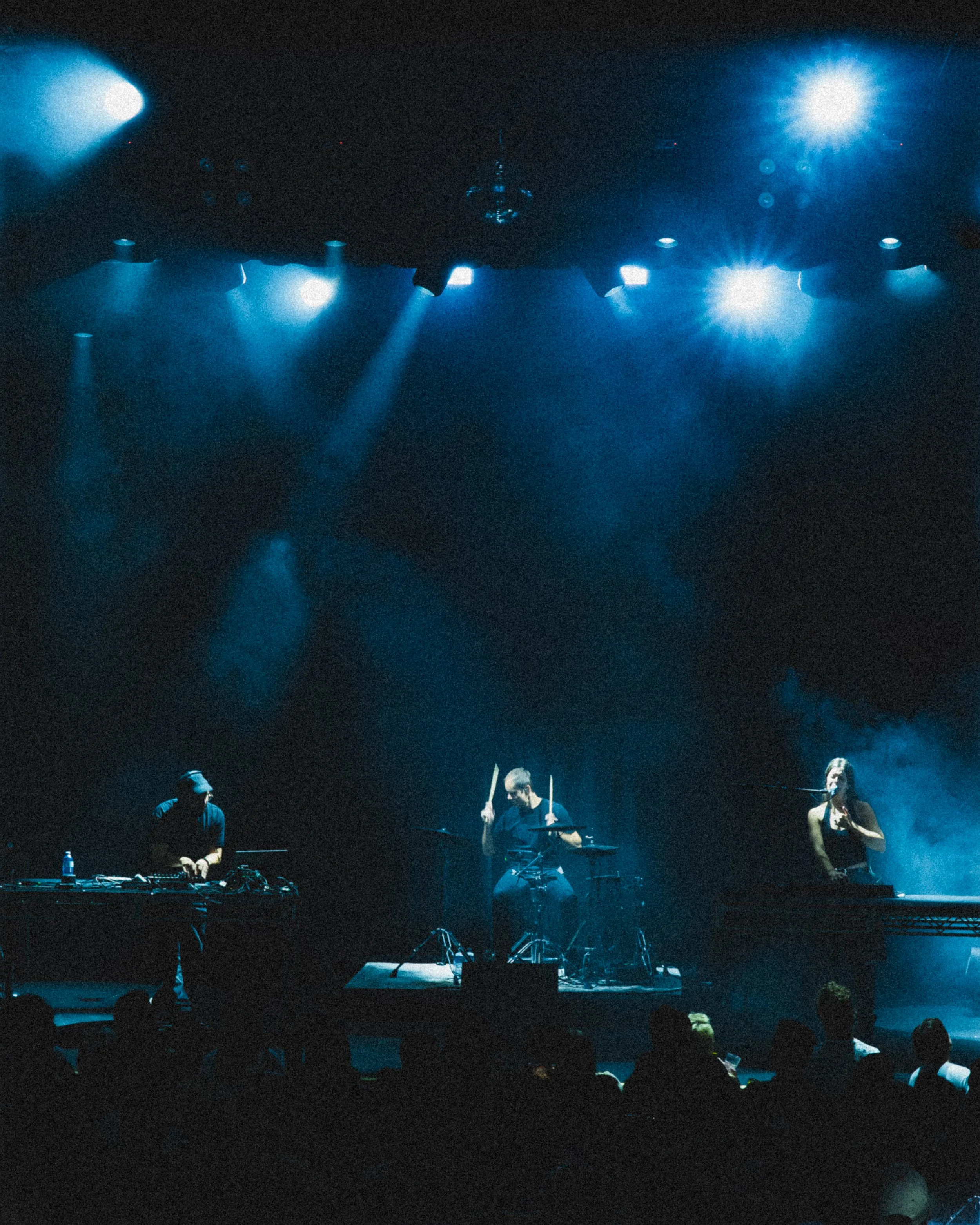 Musicians performing on stage under blue stage lights in a dark concert venue, with the audience visible in the foreground.