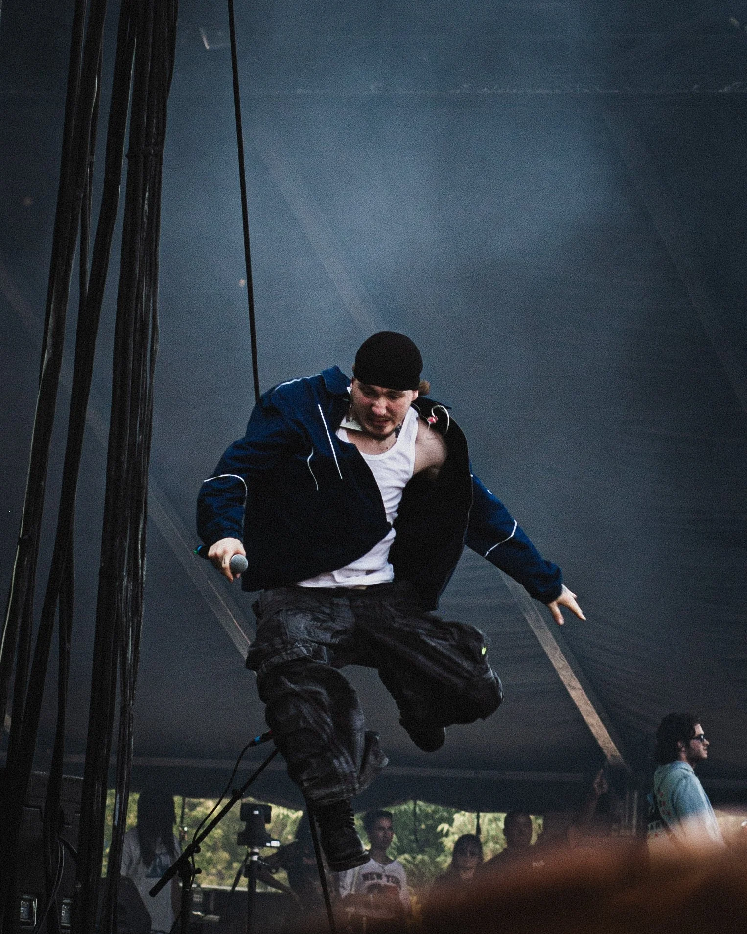 Performer jumping on stage during a concert or event, holding a microphone, wearing a black beanie, white tank top, dark jacket, and cargo pants, with audience members in the background.
