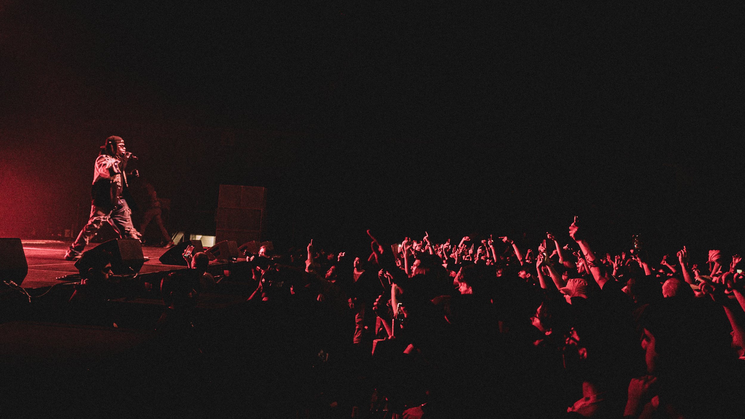 A performer on stage at night, holding a microphone, with a large audience raising their hands and phones, illuminated by dim red lighting.