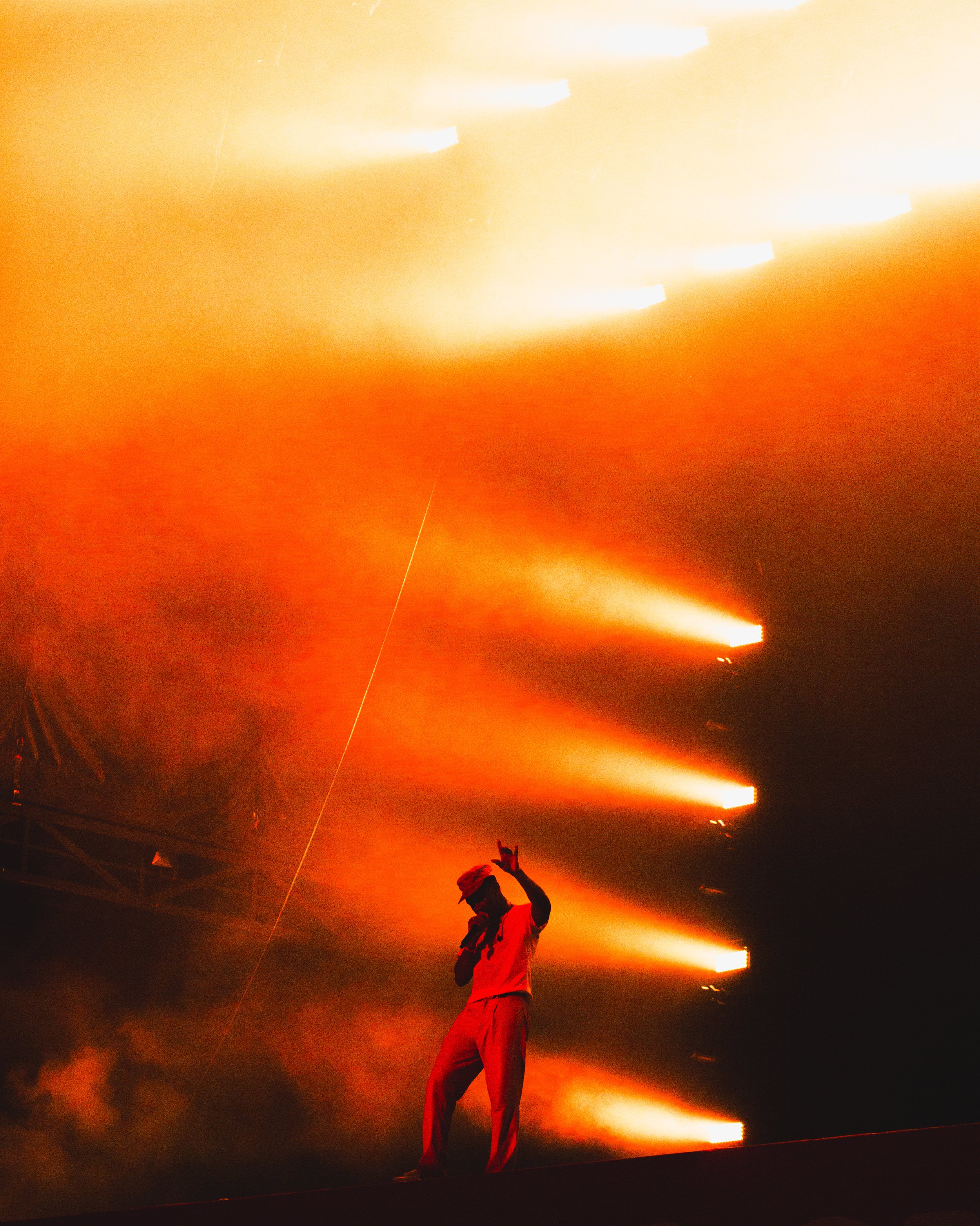 Performer on stage with bright orange and yellow lighting effects in the background, holding a microphone and making a peace sign gesture.