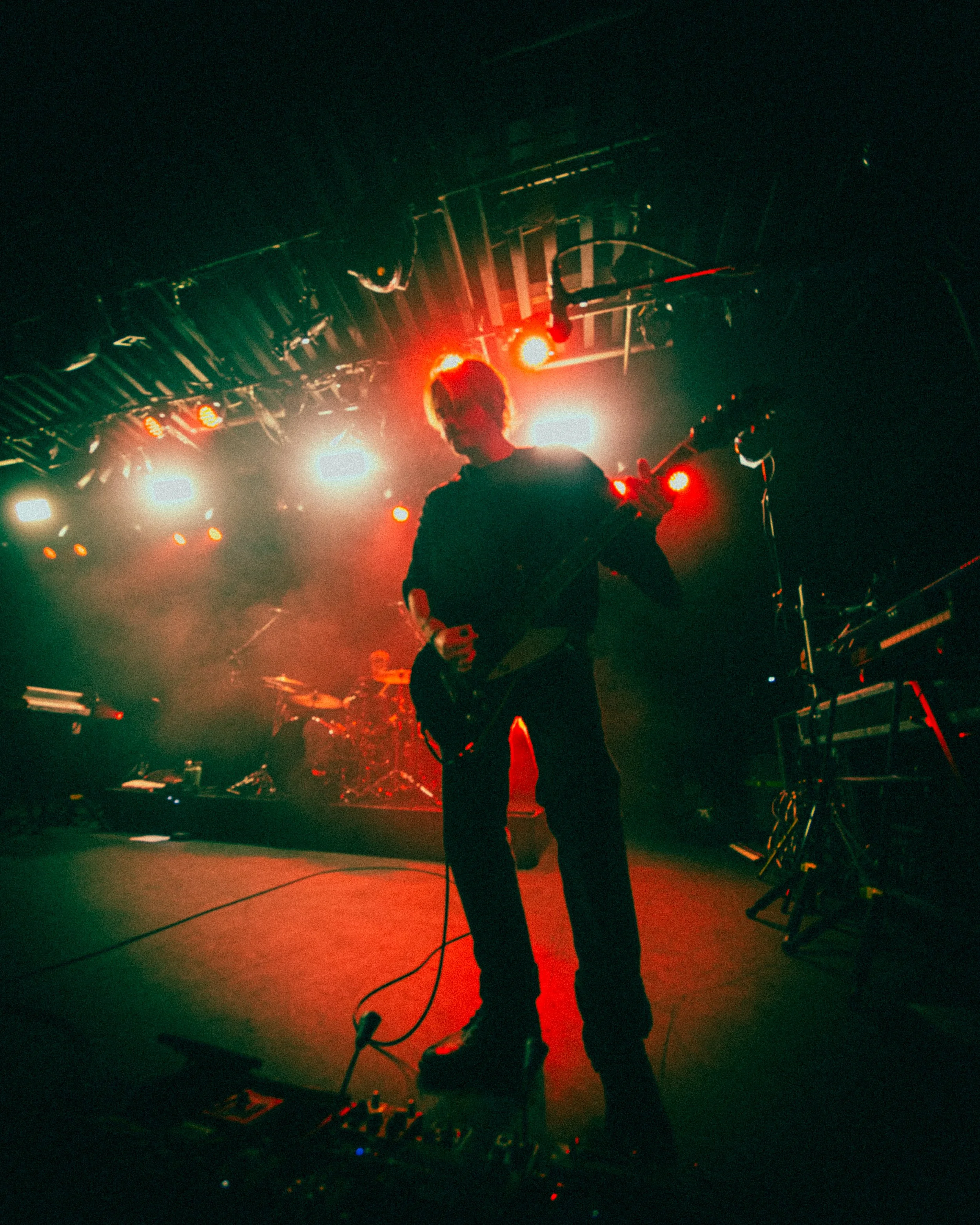 Musician playing electric guitar on a stage illuminated by red and white lights, with electronic equipment and a drum set in the background.