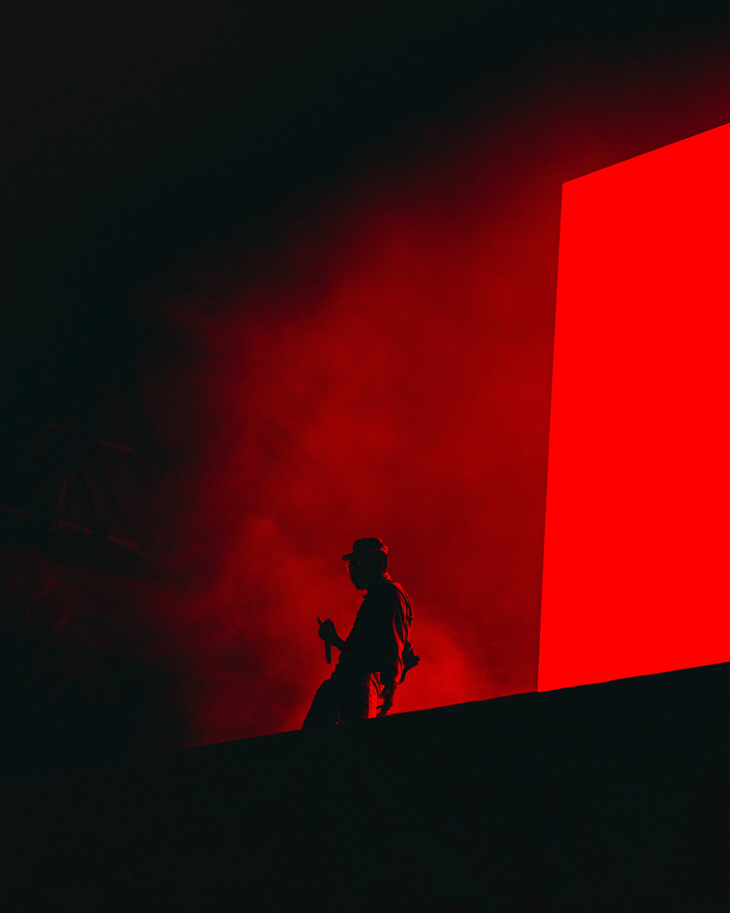 Silhouette of a person wearing a hat and glasses, holding an object, standing against a dark background with a large red illuminated structure.