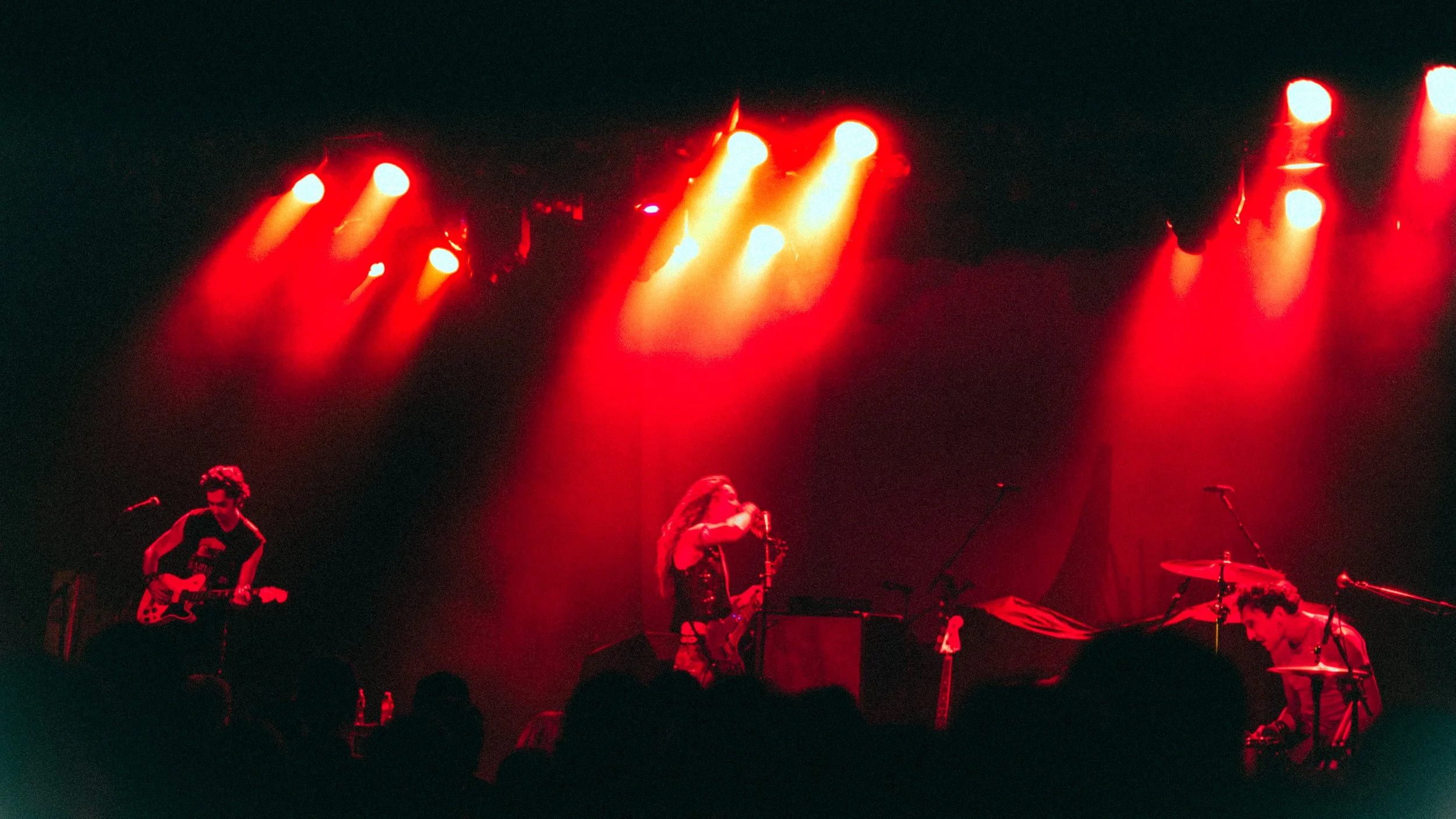 A band performing live on stage illuminated by red and orange stage lights. The band consists of a guitarist on the left, a female singer with a saxophone in the center, and a drummer on the right, with audience silhouettes in the foreground.