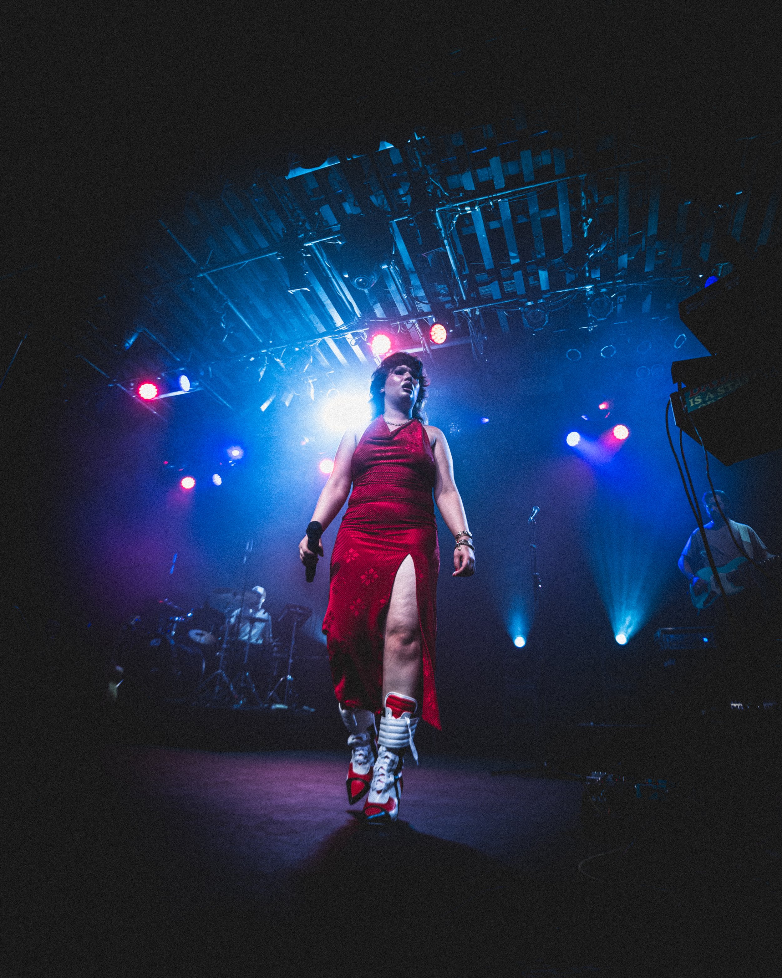 Performer in a red dress and high-top sneakers walking on stage with colorful lights, band members playing instruments in the background.