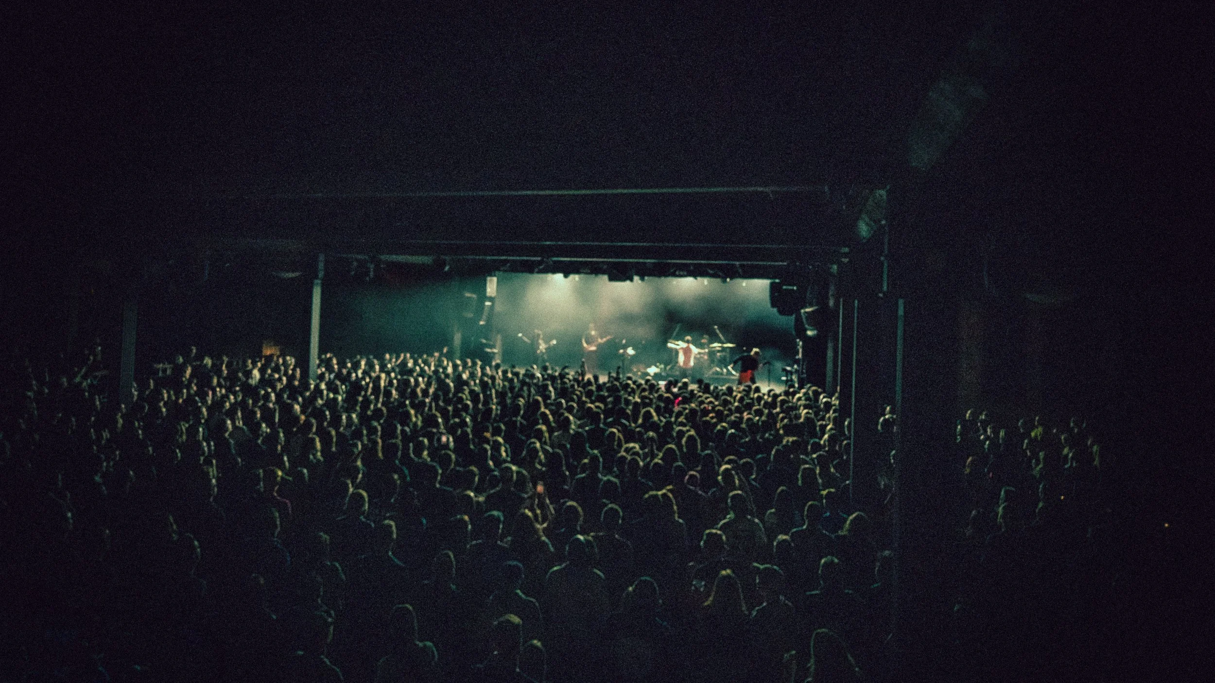 A large crowd at a concert or music event, facing a stage with performers and lighting.