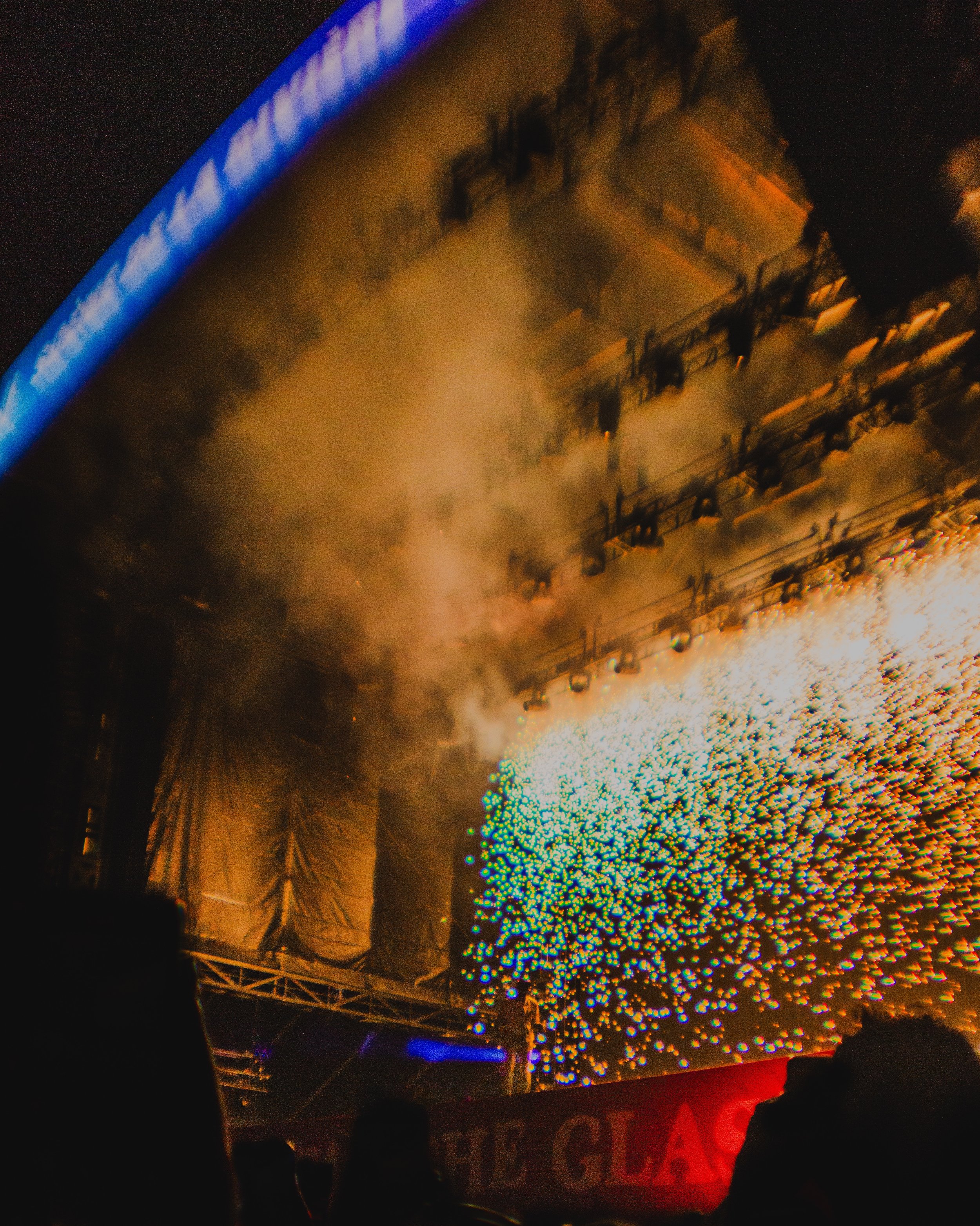 Colorful fireworks exploding over a nighttime stage with bright blue lighting and smoky haze, with silhouettes of people watching.