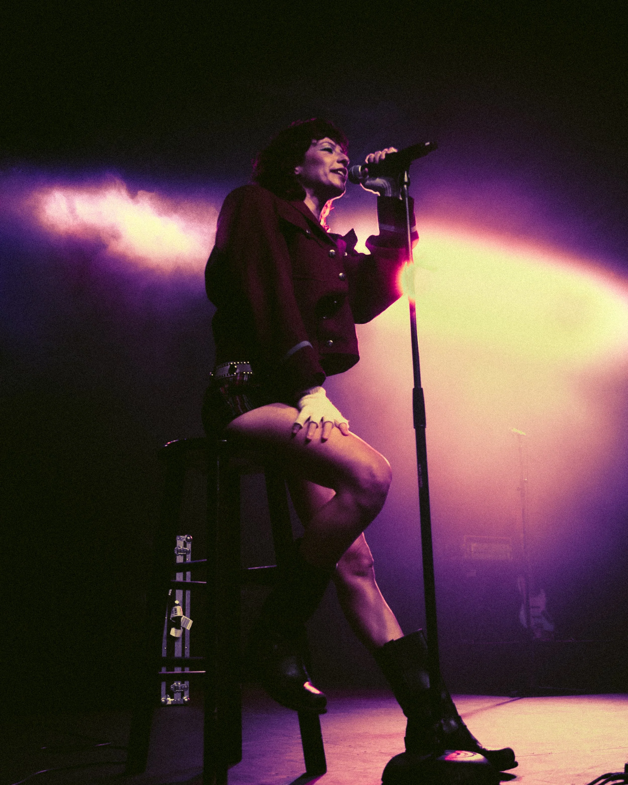 A woman singing on stage, sitting on a stool with one leg crossed over the other, under purple and yellow stage lighting.