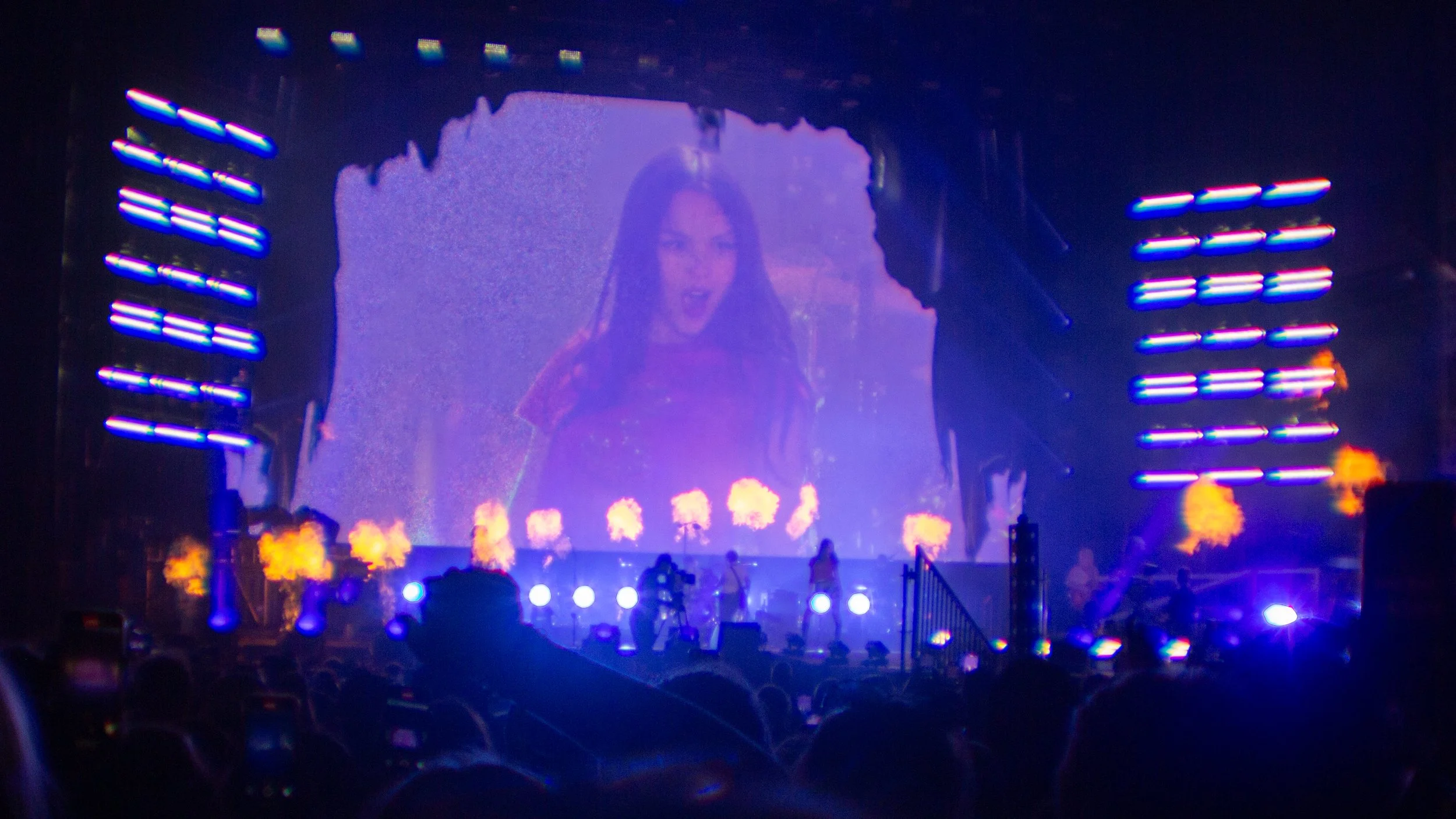 A concert stage with a large screen displaying a woman singing. Bright lights and special effects like flames are visible, with an audience in the foreground.