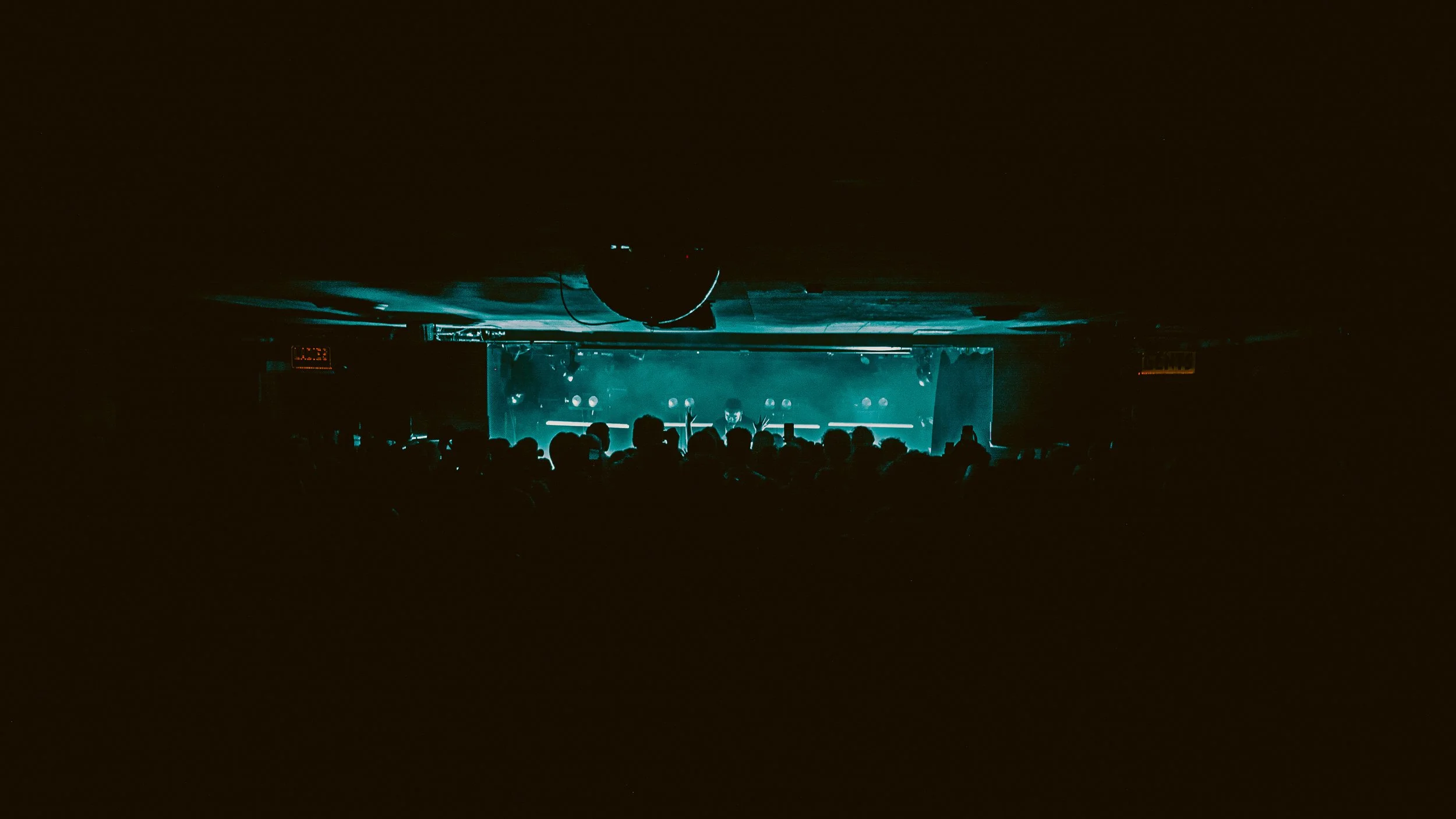 Dark concert venue with audience silhouette facing a illuminated stage with blue lighting effects.