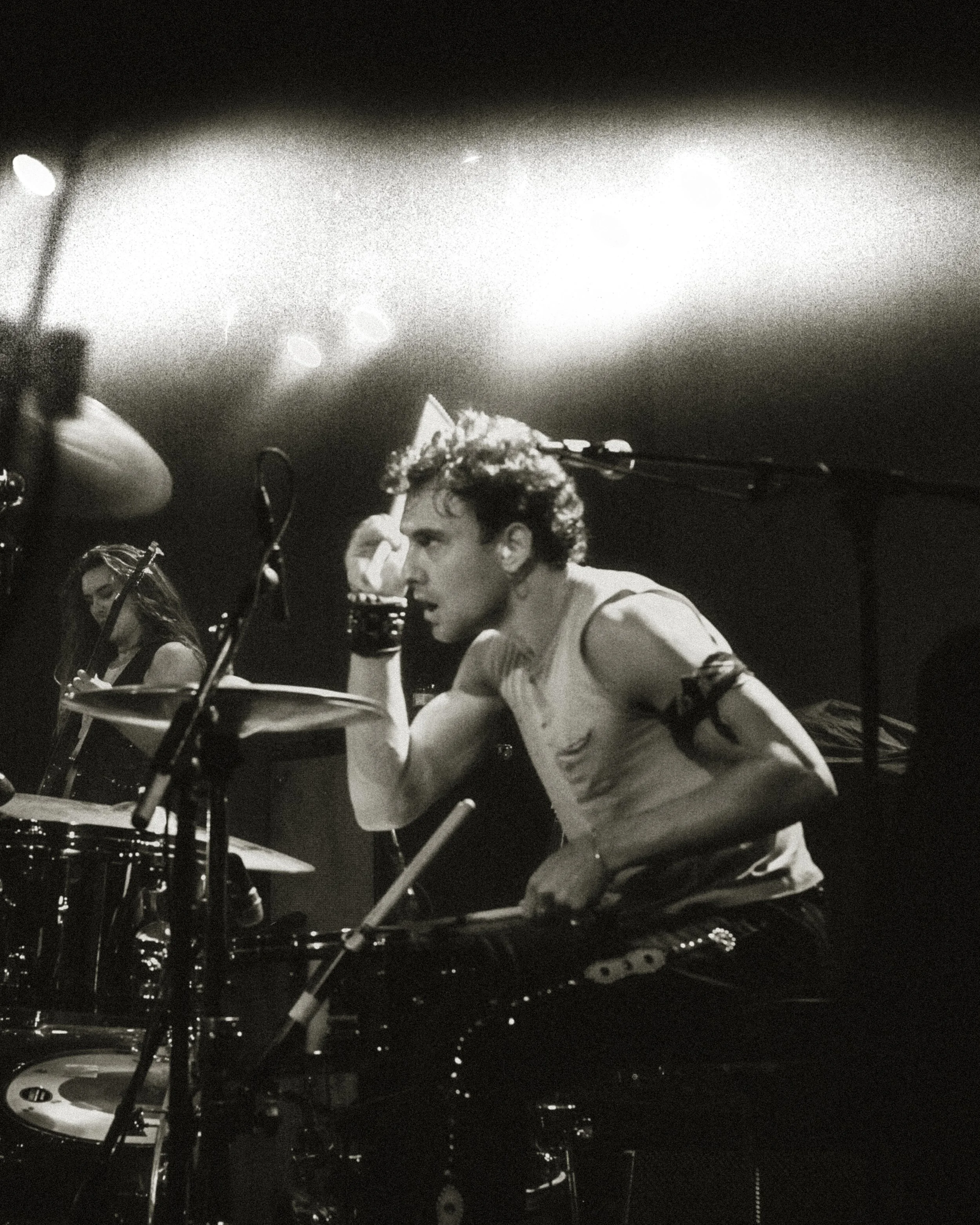 A person wearing a party hat, sleeveless shirt, and armbands, sitting at a drum set on stage, in black and white, with a woman playing guitar in the background.