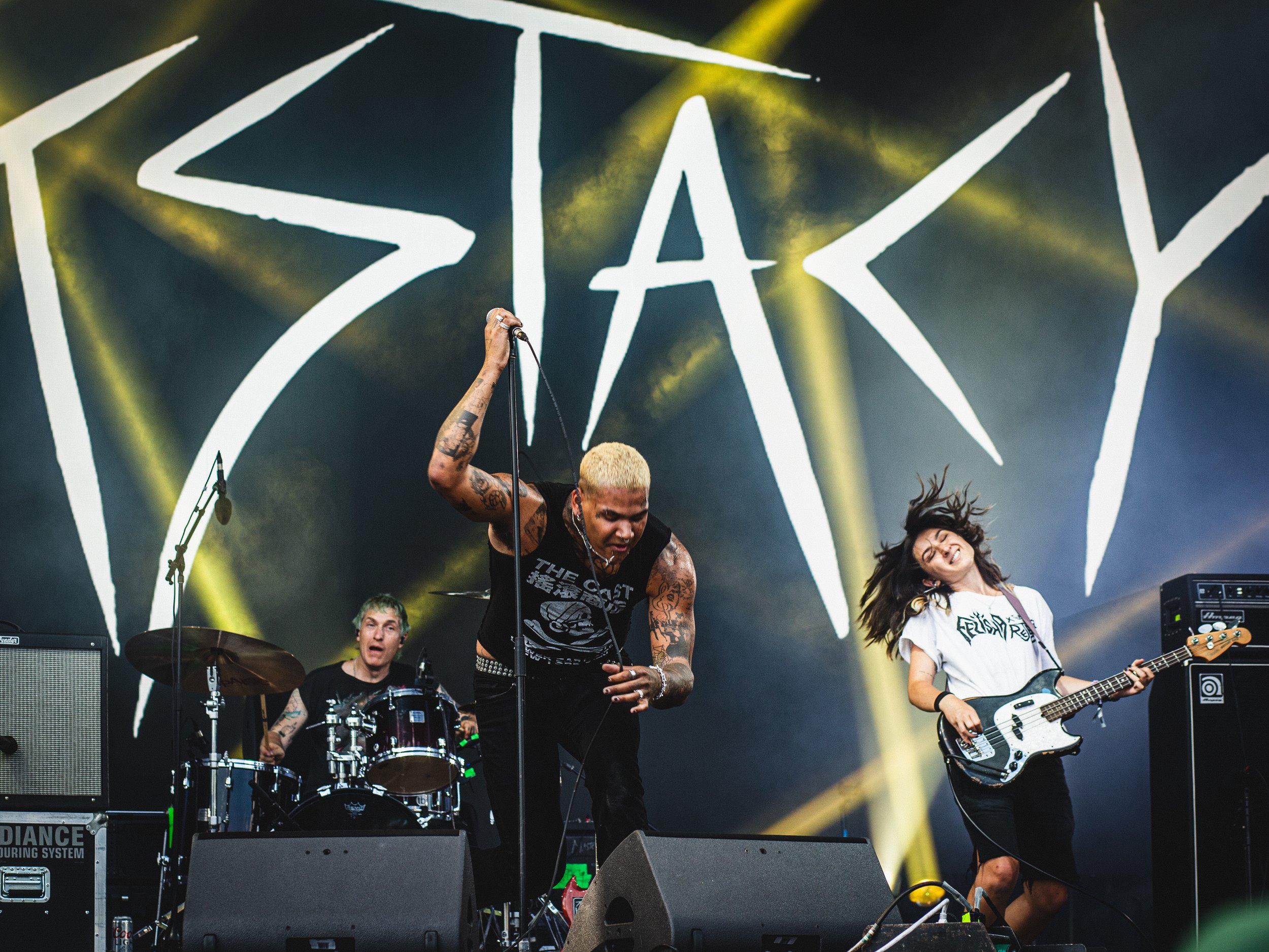 A band performing live on stage with a large logo in the background. The vocalist is holding a microphone stand, with a drummer and a bassist playing, and yellow stage lighting details.