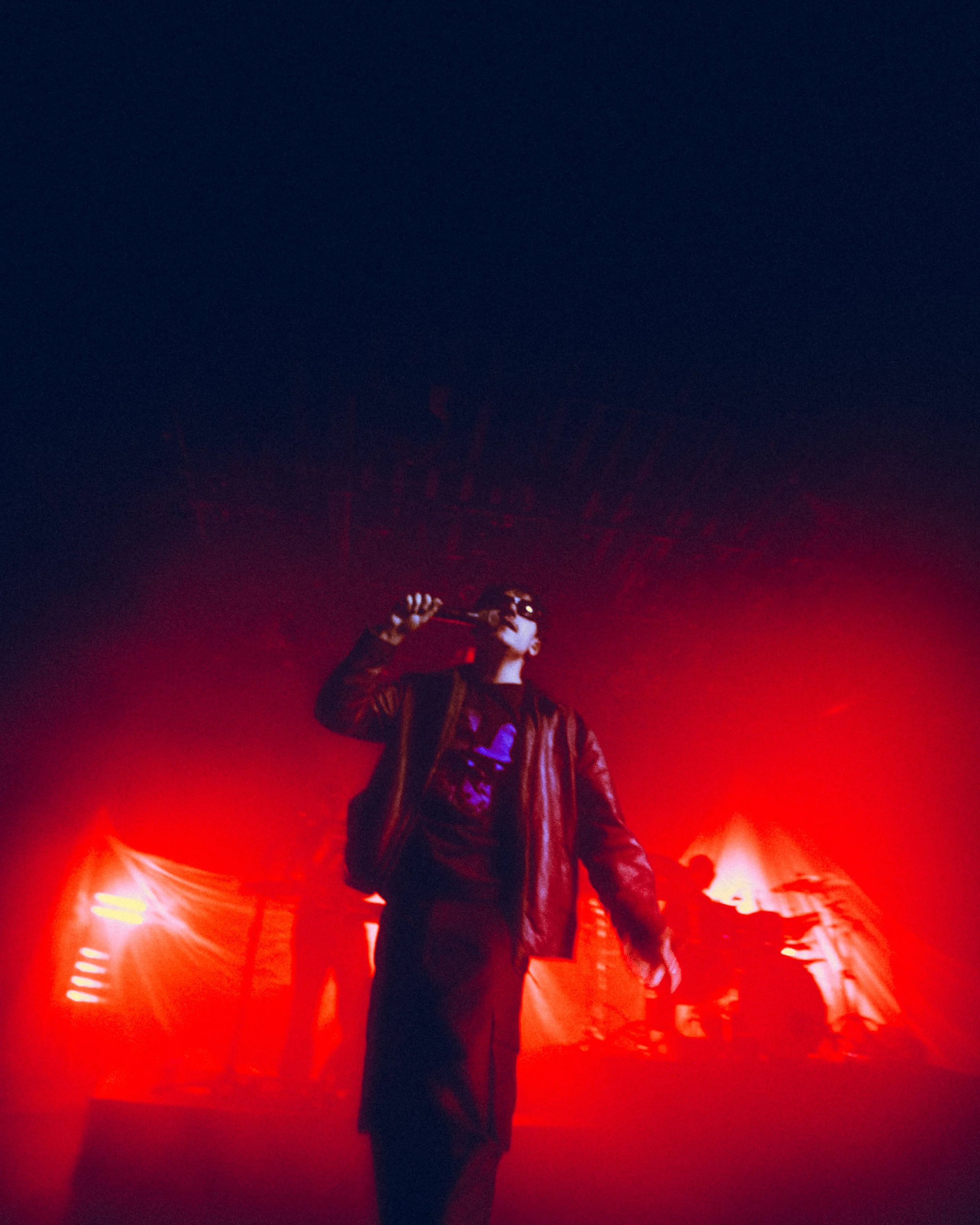 Performer on stage at night, wearing sunglasses and a leather jacket, illuminated by red stage lights, with a dark sky overhead.