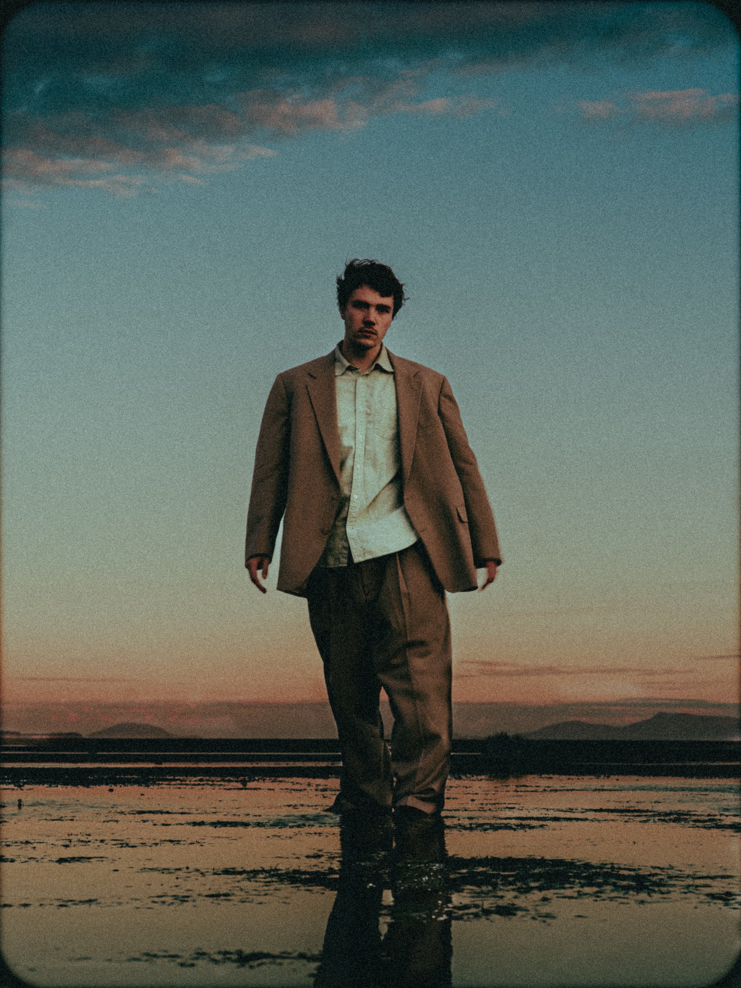 A young man in a tan suit and white shirt standing in water at sunset with mountains in the background.