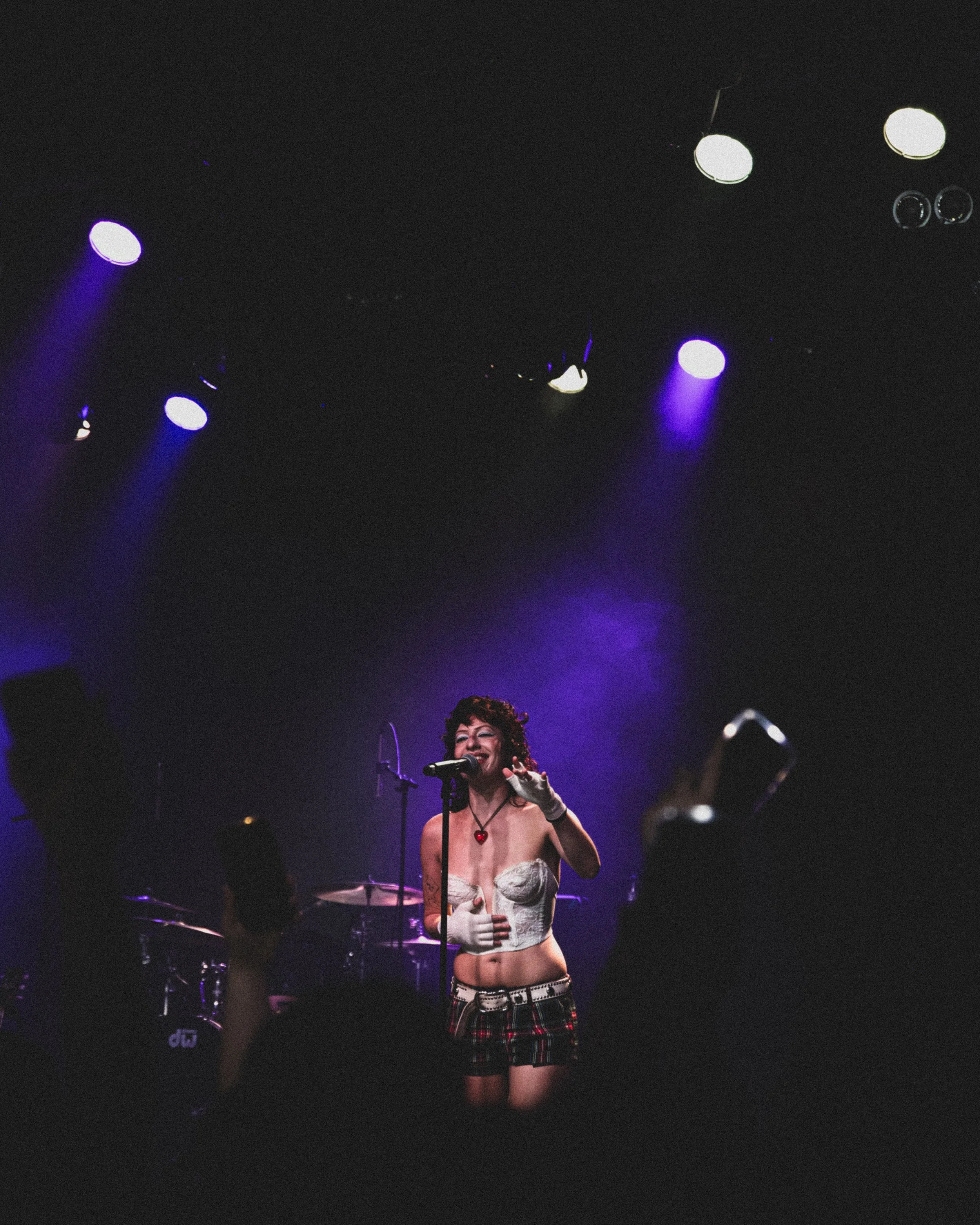 Performer singing on stage with purple lighting, wearing a white corset, plaid skirt, and white gloves, holding a microphone, with audience holding cameras