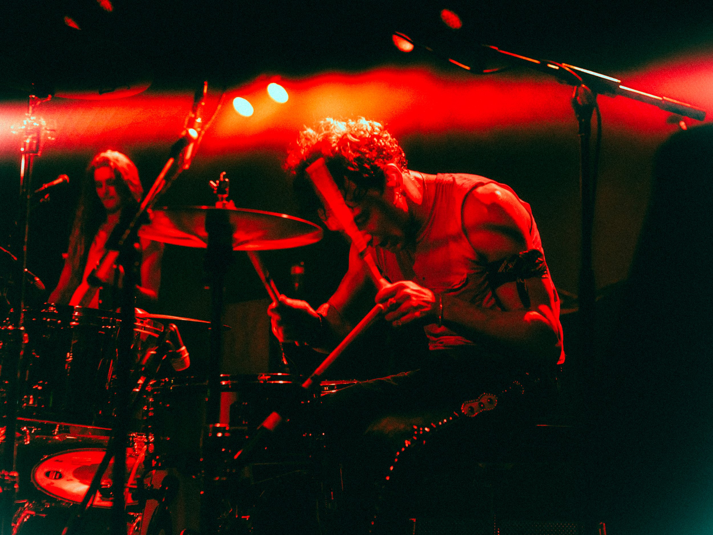 Musicians performing on stage illuminated by red lights, one playing drums and another with a microphone stand.