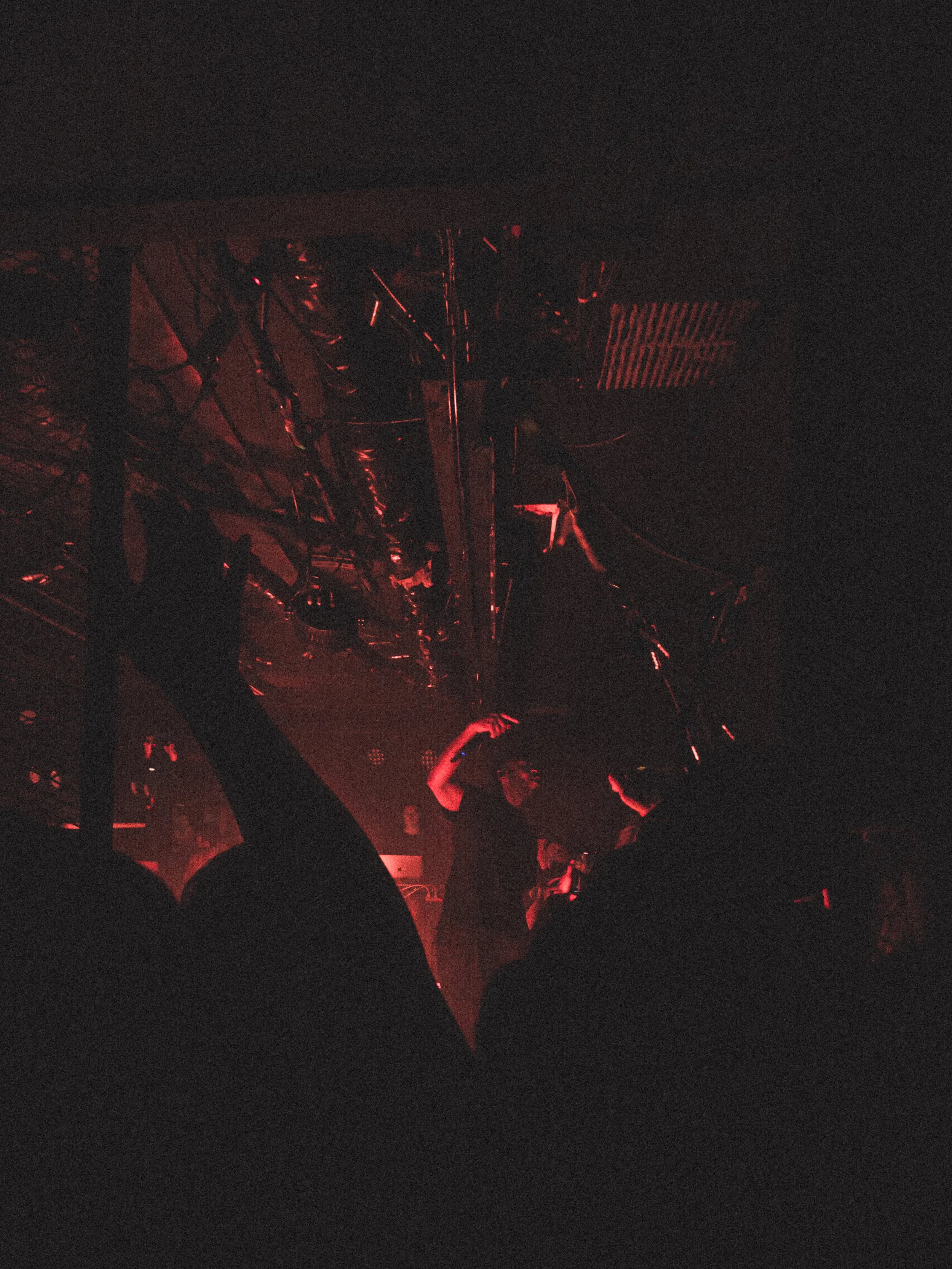 A person on stage performing in a dark, red-lit concert or club environment with metal scaffolding and equipment overhead.